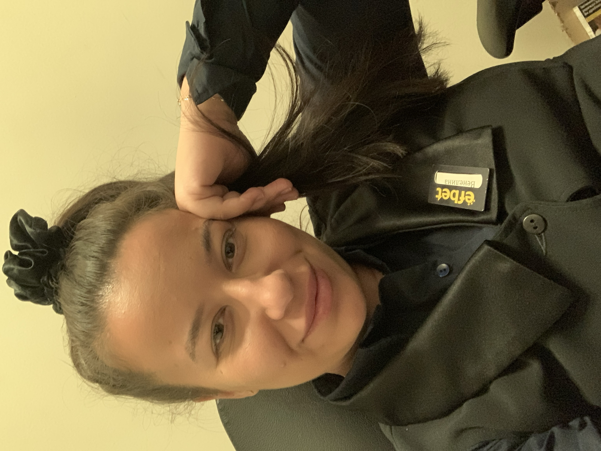 In a head-and-shoulders selfie, a woman with dark hair pulled up into a high ponytail with a scrunchie is seen resting her head on her right hand. She is wearing a black collared shirt and a black vest. A name tag, which reads "efbet Венелина", is pinned to the left side of her vest. The background is a plain, light yellow wall. The lighting suggests an indoor environment, likely an office or a similar workspace. The overall scene is casual and personal, typical of a selfie. There is no discernible information about the specific location within Burgas, Bulgaria, or any specific actions beyond the pose and attire.