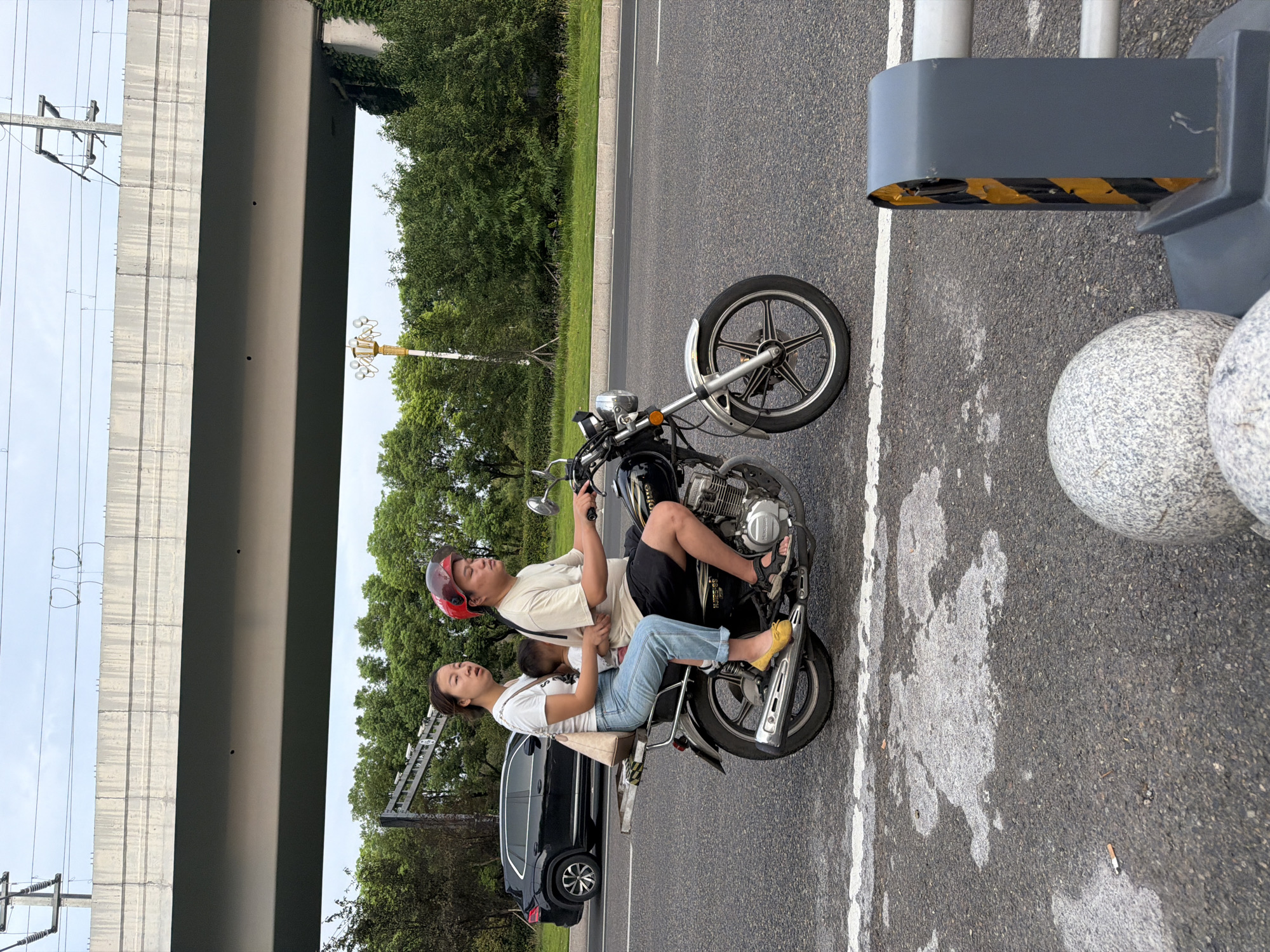 A family of three rides a black motorcycle on a paved road. The driver, a man wearing a beige t-shirt, black shorts, and a red helmet, operates the vehicle. A woman in a white t-shirt and blue jeans sits behind him, holding a young child. The child, wearing a white shirt, is nestled between the two adults. The motorcycle has a rear rack with a beige bag on it. The scene is set outdoors with a large elevated concrete structure overhead, likely an overpass or bridge. Lush green trees line the roadside, and a black SUV is visible in the background to the left. The road surface is asphalt with white lane markings. A street light with multiple lamps stands to the right of the trees. In the foreground, a gray traffic barrier with yellow and black hazard stripes is present, along with two large, speckled gray spherical objects. The overall impression is a typical urban or suburban scene. The presence of the street lamp with multiple lamps is a characteristic feature of certain urban areas in China, and the style of the motorcycle and the clothing of the individuals suggest an Asian setting. This particular scene is located in Jiangjin District, Chongqing, China.