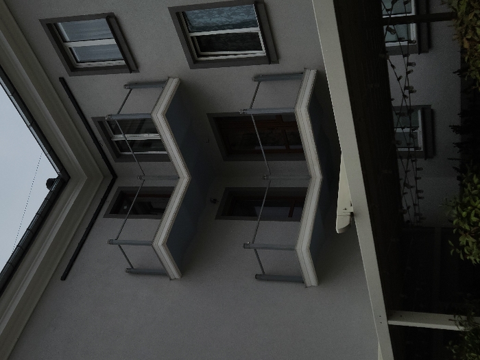An exterior view of a grey-colored building in Krasnodar, Russia, under an overcast sky is depicted. The primary focus is a prominent architectural corner featuring two stacked, L-shaped balconies. Each balcony exhibits a white underside trim, a light blue soffit, and modern railings constructed from grey metal posts, a top rail, and clear glass infill panels. Behind these balconies are dark-framed glass doors. To the right of the corner, two vertically aligned rectangular windows with white frames and dark grey surrounds are visible; the upper window contains a white curtain, while the lower displays a patterned sheer curtain. A light grey/white awning or pergola structure, featuring a visible retractable mechanism, extends from the building below the lower balcony. Beneath this structure, string lights are visible, and green foliage occupies the bottom foreground. No individuals or ongoing actions are present.