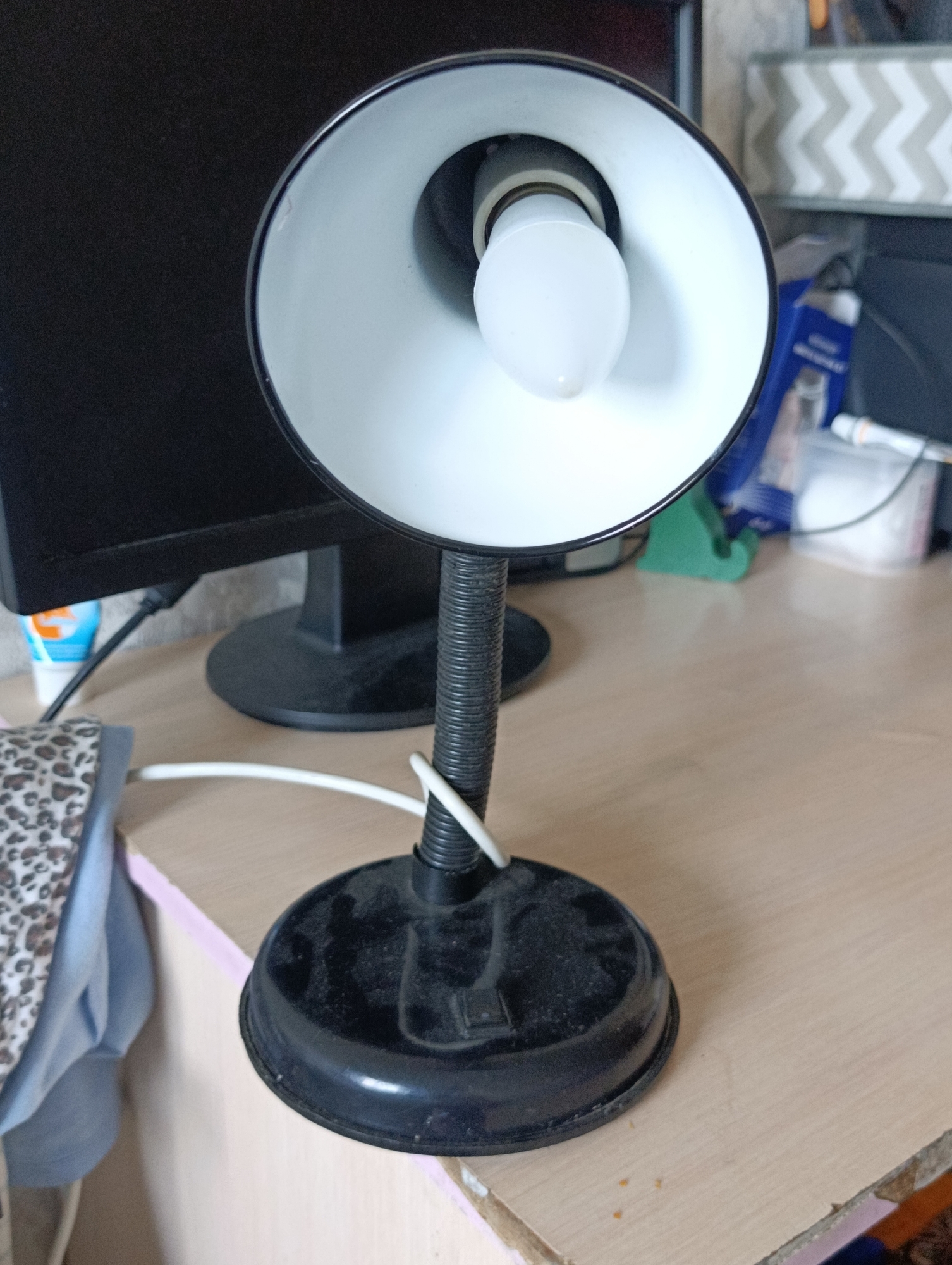 The image shows a black desk lamp with a white interior shade and a visible LED light bulb. The lamp has a flexible gooseneck and a circular base. The lamp is positioned on a light-colored wooden desk, which appears to be cluttered with various objects in the background, including a computer monitor, a green object, and some containers. To the left of the lamp, a patterned fabric, possibly clothing or a bag, is visible. The scene is indoors, likely a home or office setting in the city of Semey, Kazakhstan.