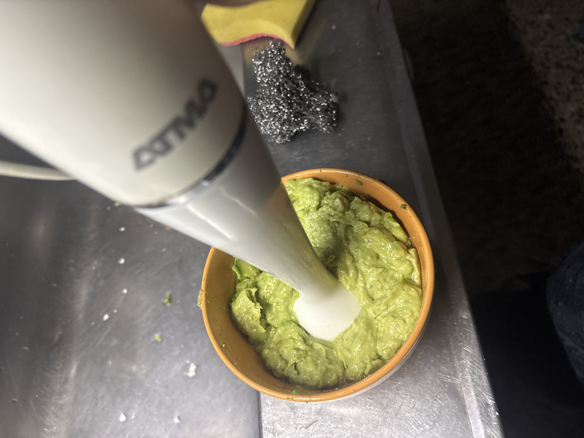A white immersion blender, with the brand name "ATMA" visible on its side, is being used to blend a bright green mixture in a small, orange-rimmed bowl. The bowl is sitting on a stainless steel surface, possibly a kitchen sink. Next to the bowl and blender is a metal scouring pad and a yellow sponge. The scene appears to be in a kitchen. The location in the city of Buenos Aires, Argentina is not discernible from the image.