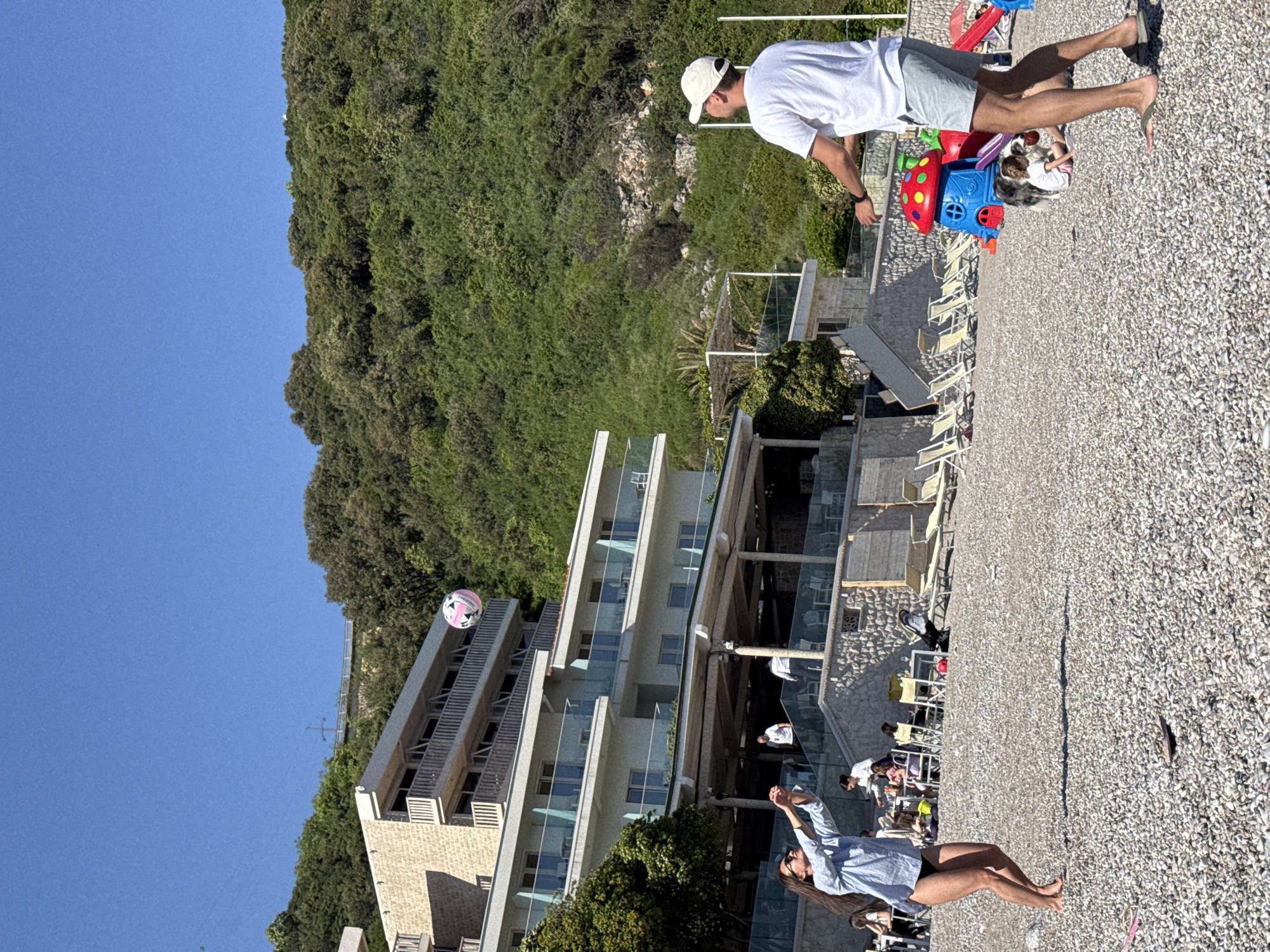 Here is a concise description of the image:
The image shows a coastal scene, likely in Prijevor, Montenegro, featuring a beachfront area with a pebble beach and a multi-story modern building with glass balconies. A woman stands on the beach, appearing to play with a pink and white ball in the air. In the background, on an elevated portion of the beach, a man stands near a child's toy car with children in it. The vegetation is lush, and the sky is clear blue. Beach chairs and other people are visible, suggesting a recreation or vacation setting.