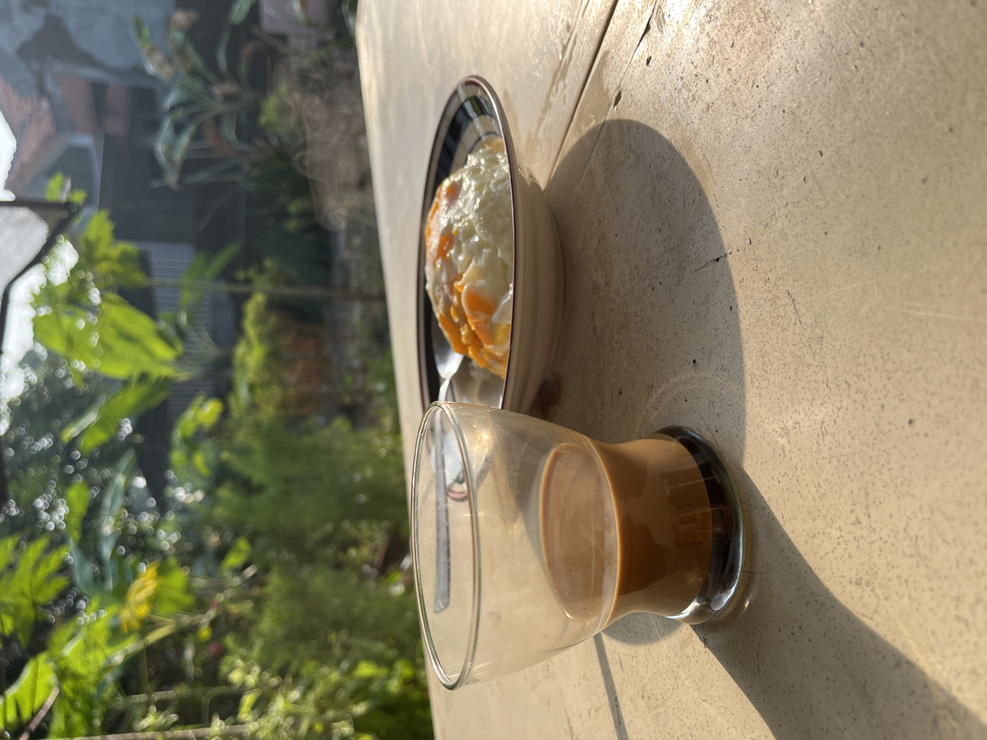 A glass of coffee and a bowl of fried eggs are on a table. The table is made of stone and is located outdoors. In the background, there are trees and houses. The sun is shining, creating a shadow from the glass on the table. The scene appears to be a breakfast setting.