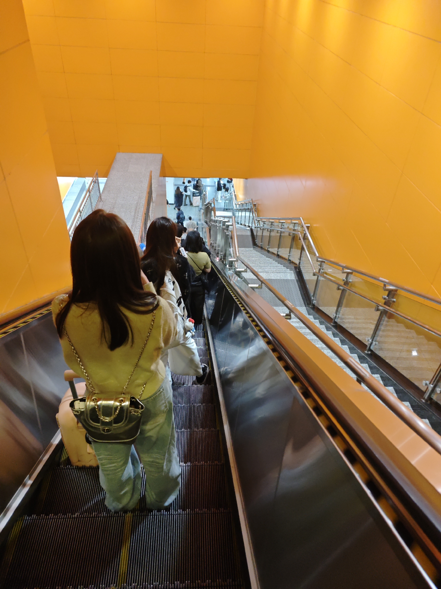 The photograph shows the view from the top of an escalator descending into a space with orange walls, likely a transportation hub or public space in Liangjiang, China. At least 4 people, women, can be seen on the escalator, with the woman in the foreground wearing a light yellow jacket and light blue jeans with a silver purse. Other people are further down, in what looks like a waiting area. The escalator is positioned alongside a stairway. The architectural design includes orange tiled walls. The lighting is artificial.