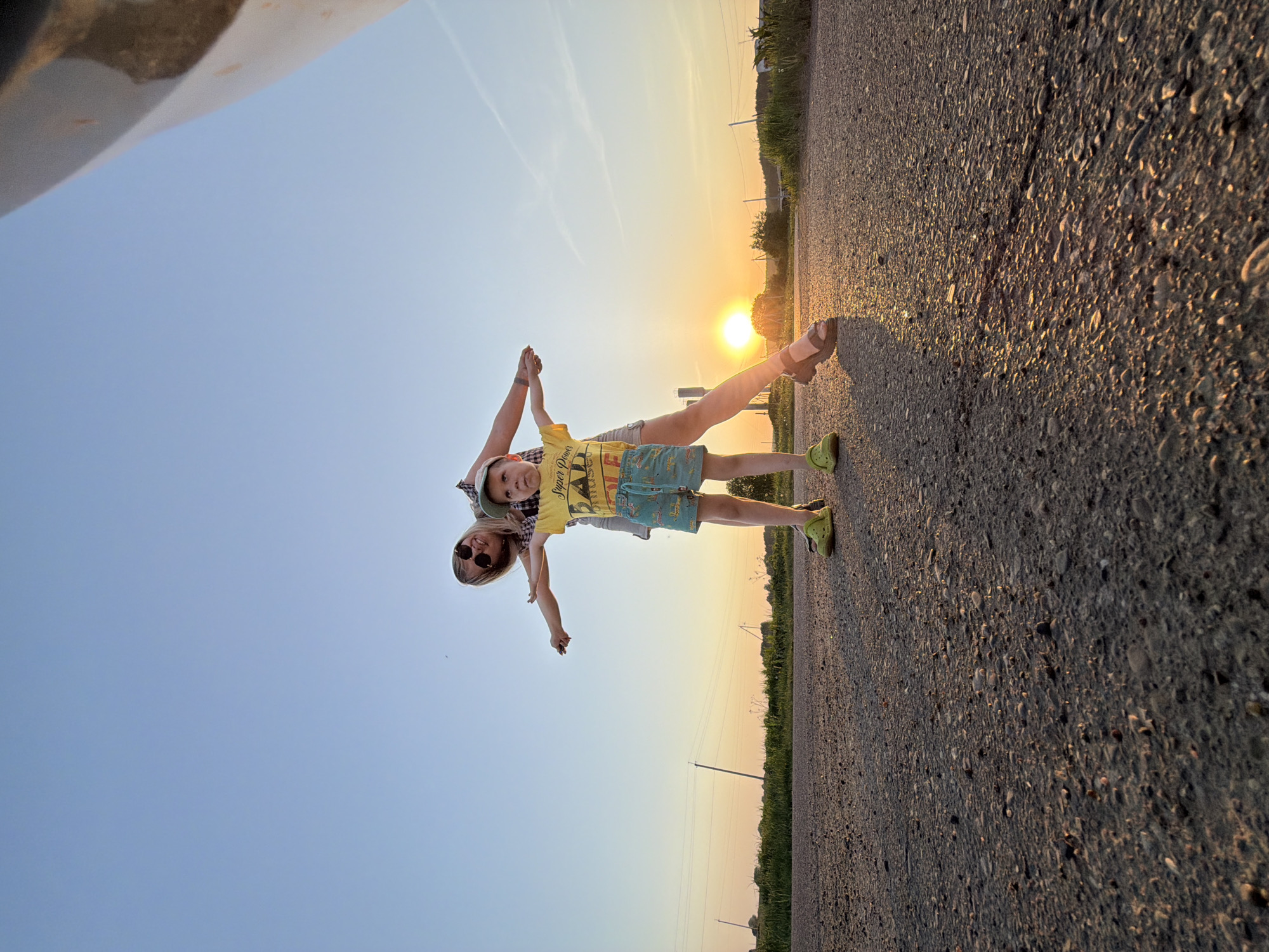 A woman and child are holding hands and laying perpendicular on what appears to be an asphalt road. The woman is wearing a plaid shirt and sunglasses. The child is wearing a yellow t-shirt, blue shorts, a hat, and green shoes. The sun is setting in the background. The photograph appears to have been taken on a road in a rural area near Чумна, Russia.
