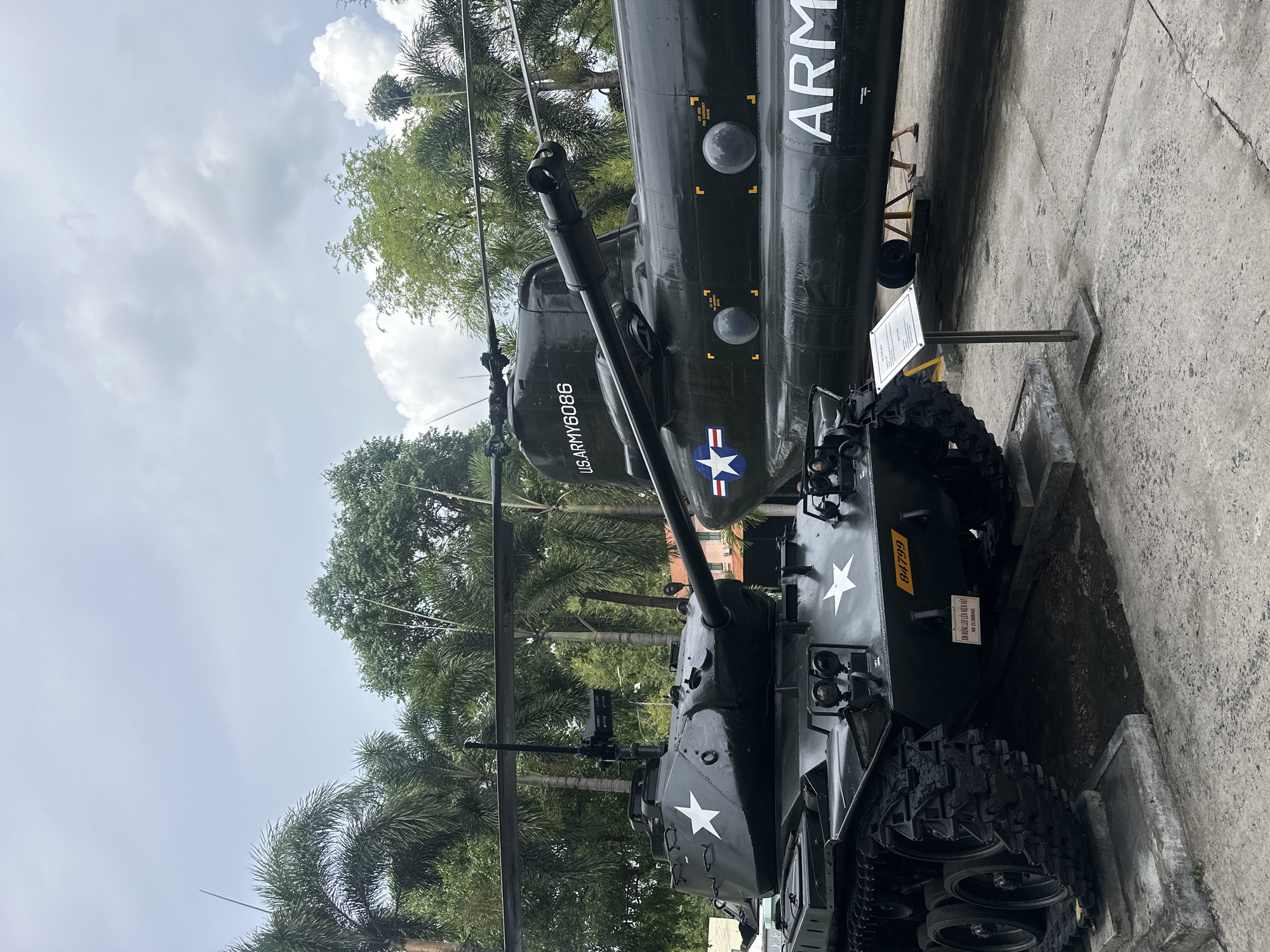 This outdoor scene shows a static display of military vehicles in Thủ Đức, Vietnam. The vehicles consist of a U.S. Army helicopter and a tank, painted dark green with white stars. The helicopter has "ARMY" printed on its side along with the text "US ARMY 6086" on the nose. The tank is directly below the helicopter. The background includes trees and a cloudy sky. The display is set on a concrete surface.
