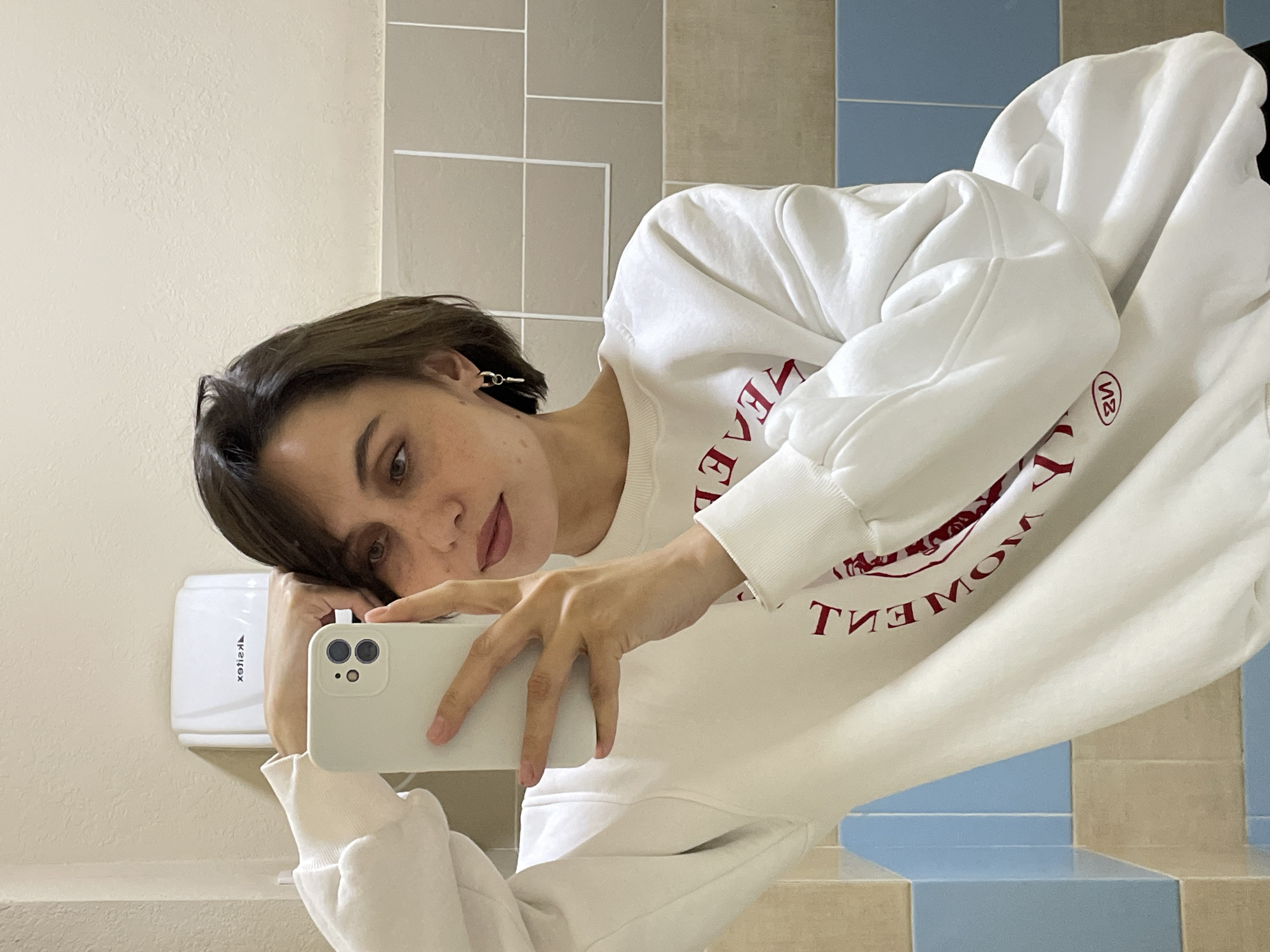 A young woman with short, dark brown hair and fair skin is depicted in a mirror selfie, taken indoors. She wears a white sweatshirt adorned with mirrored red text, which appears to read "LIVE THE MOMENT" and includes a small circular "VS" or "SV" logo. Her left arm and head are resting on a white, counter-like surface, while she holds a light-colored smartphone with a multi-lens camera in her right hand to capture the image. A subtle lip tint is visible on her lips, and she has a small earring in her left ear.
The background consists of tiled walls, featuring light beige squares in the upper section and a combination of light blue and beige tiles below. To her left, a white wall-mounted hand dryer with the mirrored brand name "Xsilex" is visible. The woman's gaze is directed towards the camera with a calm, somewhat pensive expression. The setting strongly suggests a public or private bathroom, in Izhevsk, Russia.