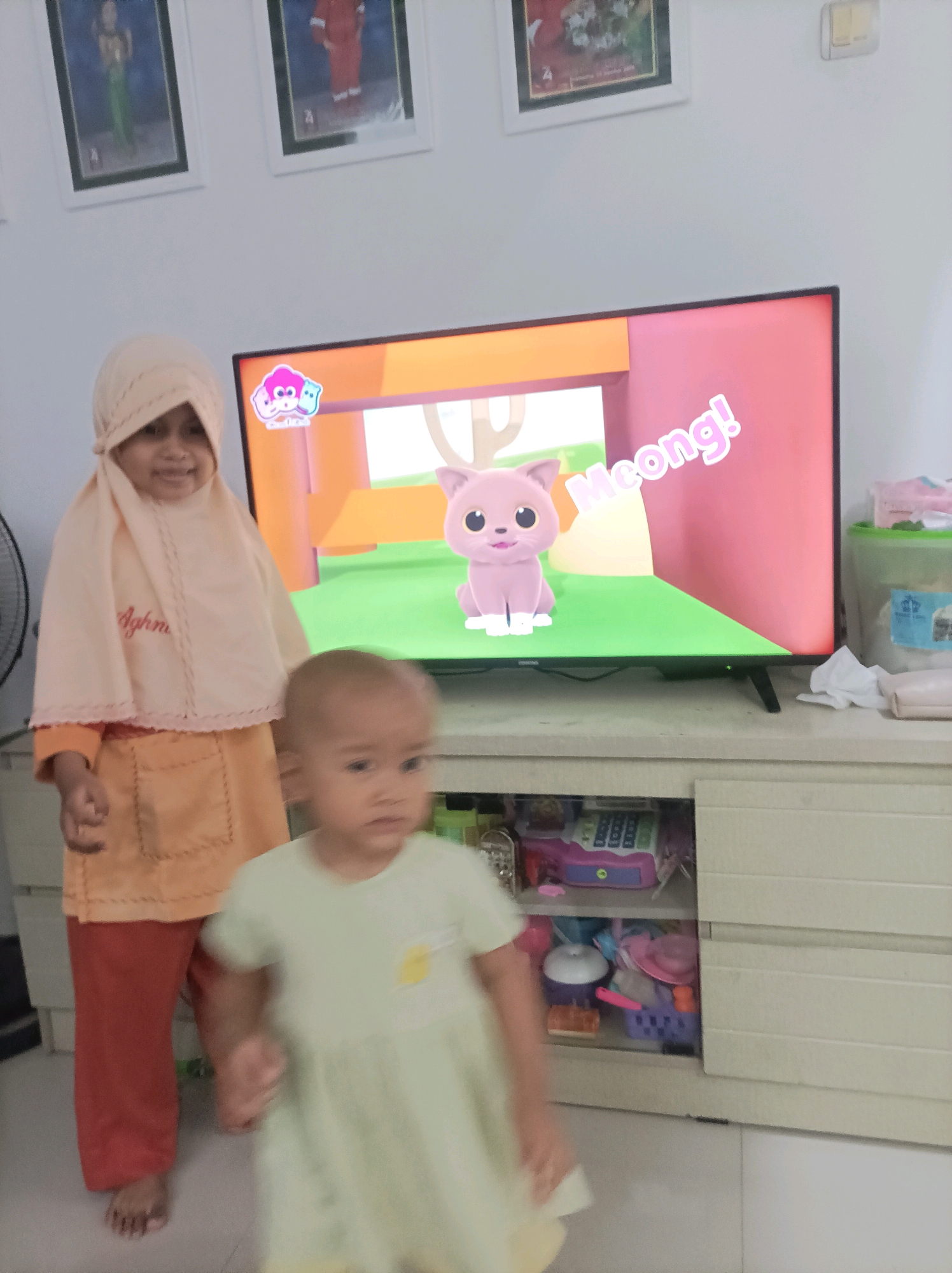 Two young children, one wearing a hijab and an orange outfit, and another in a light green dress, are standing in front of a television screen displaying a cartoon of a pink cat with the word "Meong!" written in large letters. The television is placed on top of a light-colored cabinet filled with various items, including toys and what appears to be a calculator. The room is illuminated, and the overall environment suggests a domestic setting in Brondong, Indonesia. The child in the hijab is looking towards the camera, while the child in the green dress is facing away, slightly out of focus.