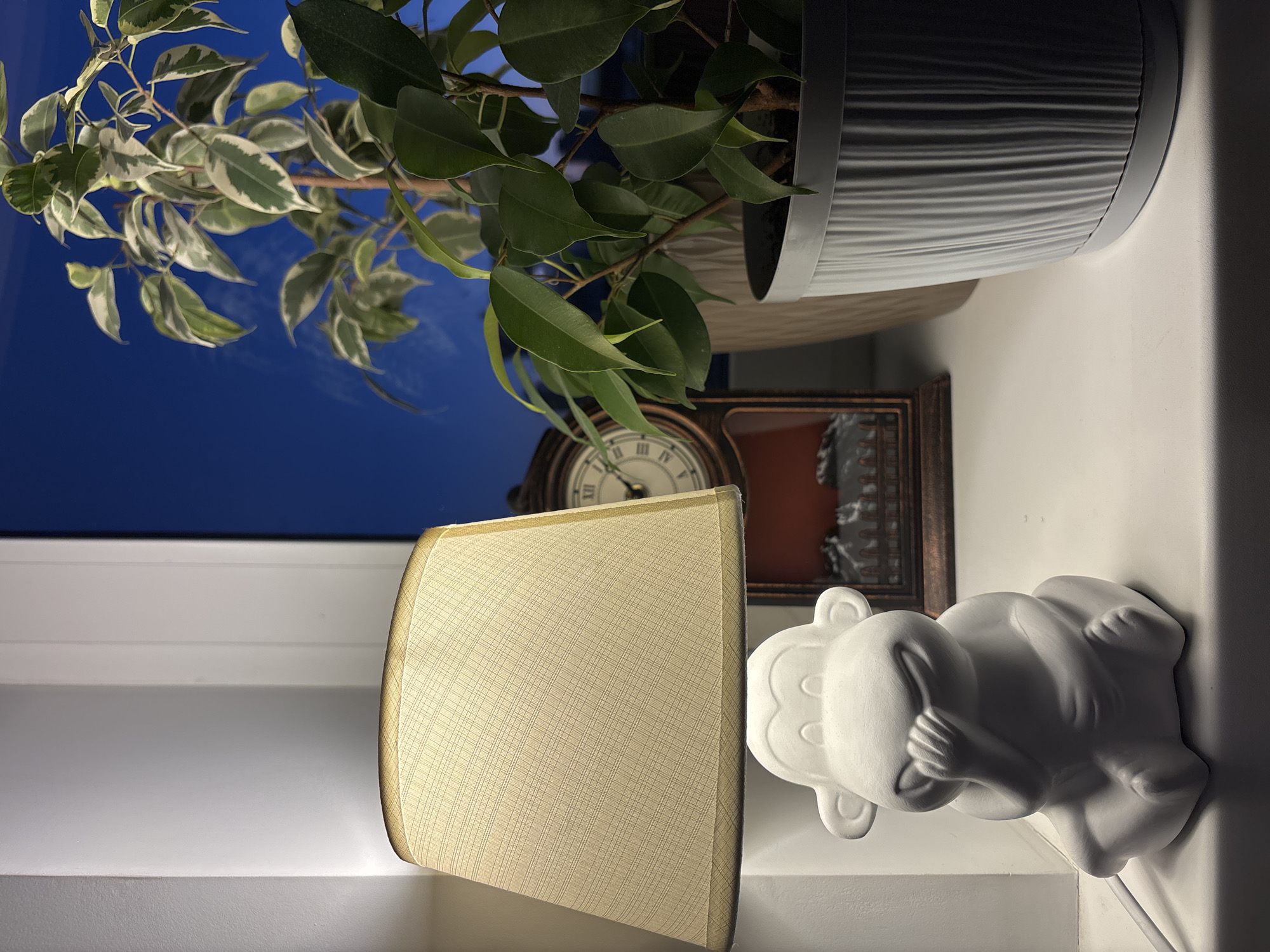 An indoor scene at night with a view of the night sky through a window. On a white windowsill, a white ceramic monkey-shaped lamp is positioned to the left. The lamp has a beige fabric lampshade. To the right of the lamp, there is a vintage-style mantel clock with a Roman numeral face. Further to the right, there is a potted plant with variegated green and white leaves. The plant is in a grey ribbed pot. The overall scene suggests a cozy indoor setting at dusk or night.