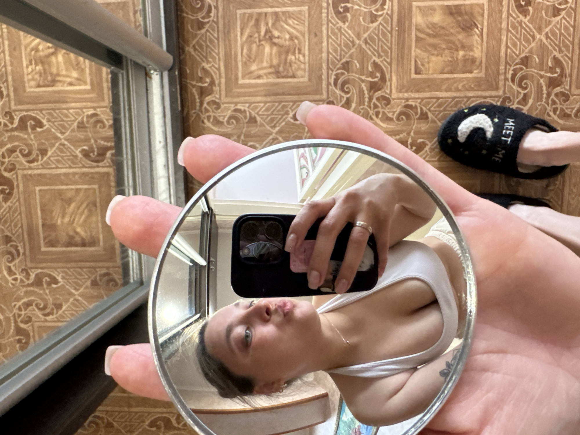 A young woman is taking a selfie in a small circular mirror that she is holding in her left hand. The mirror reflects her face, which is contorted into a pout, her right arm, and the top of her chest. She is wearing a white tank top. She has a ring on her left ring finger. Her nails are long and polished. Behind her, on the floor, is a black slipper with a crescent moon design and the words "MEET ME" embroidered on it. The floor appears to be tiled with a brown pattern. A metal frame, possibly a window or door frame, is visible to the left of the frame, with some light reflecting off of it. The overall scene suggests a casual indoor setting. There is no indication of time of day or weather.