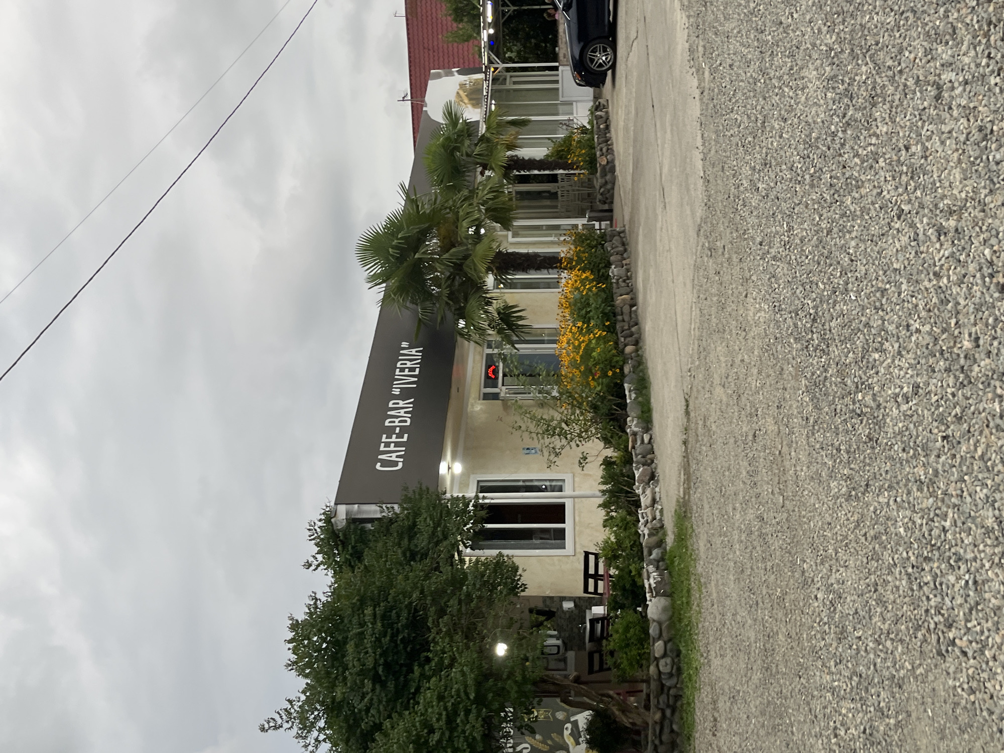 A cafe-bar named "IVERIA" is depicted in a low-angle shot under an overcast sky. The building has a light-colored facade and a dark awning with the name of the establishment printed in white lettering. In front of the cafe, there are several palm trees and a flower bed with yellow blooms. A stone wall borders the flower bed. To the left of the cafe, there is a tree with dense foliage, and a small seating area with tables and chairs. A dark-colored sedan is parked on the paved area to the right of the cafe. The foreground is covered in gravel. The scene appears to be in a rural or suburban area, possibly in the city of ღრმაღელის თემი, Georgia.