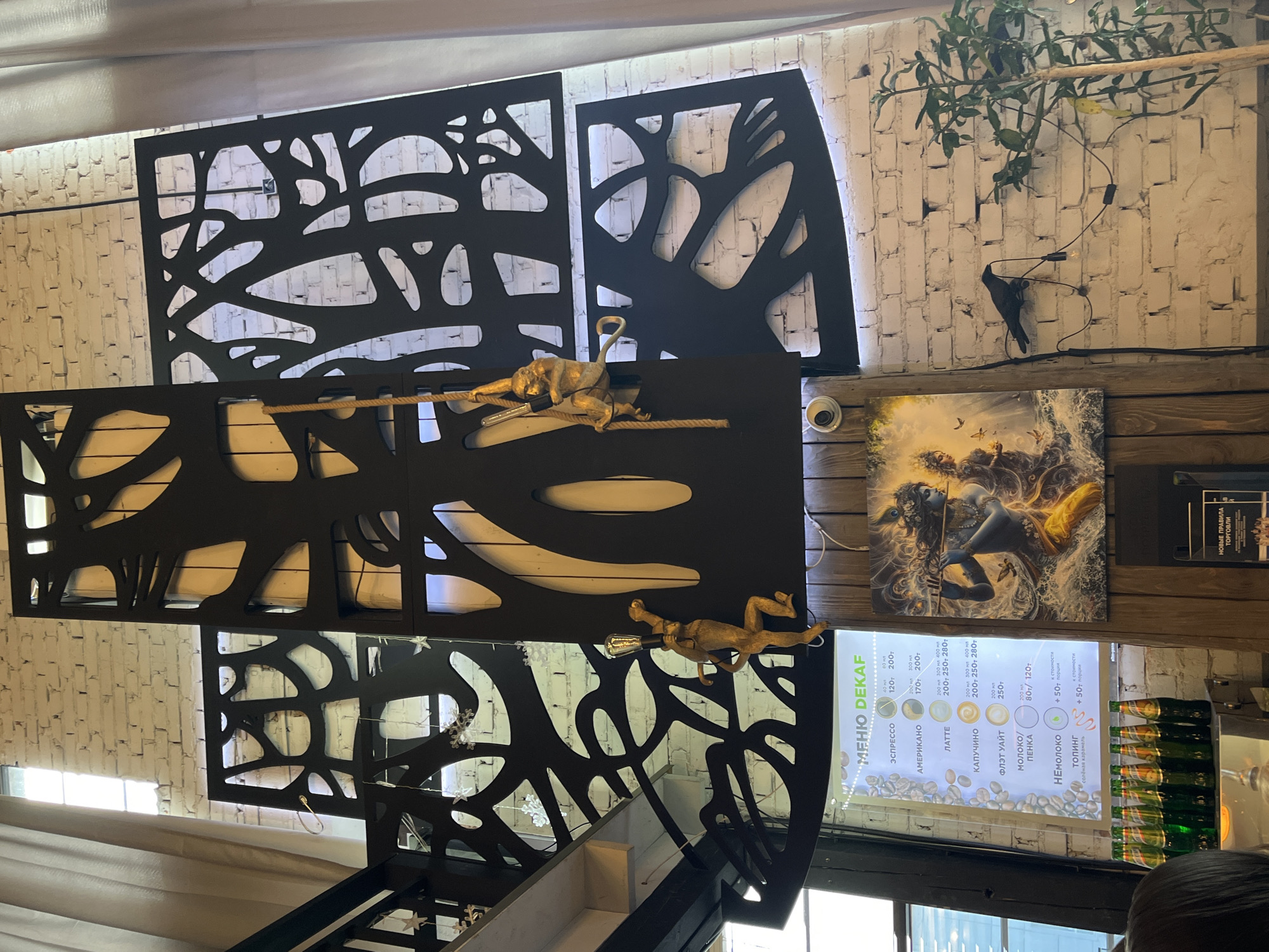 The image depicts the interior of a cafe in Moscow, Russia. A large, black, abstract metal screen with organic, tree-like cutouts dominates the upper portion of the scene. Two decorative golden monkey figures are attached to the screen, one climbing a rope holding a lightbulb, and the other in a dynamic pose. In the lower half of the image, a menu board labeled "МЕНЮ DEKAF" is visible, listing various coffee drinks with prices. To the right of the menu, a vibrant painting featuring a deity-like figure playing a flute is displayed on a wooden panel. A black crow sculpture is mounted on the white brick wall to the right of the painting, with a cord and lightbulb extending from it. Several green glass bottles are lined up on a counter in the lower left corner. The environment appears to be an indoor space with white brick walls and ambient lighting.