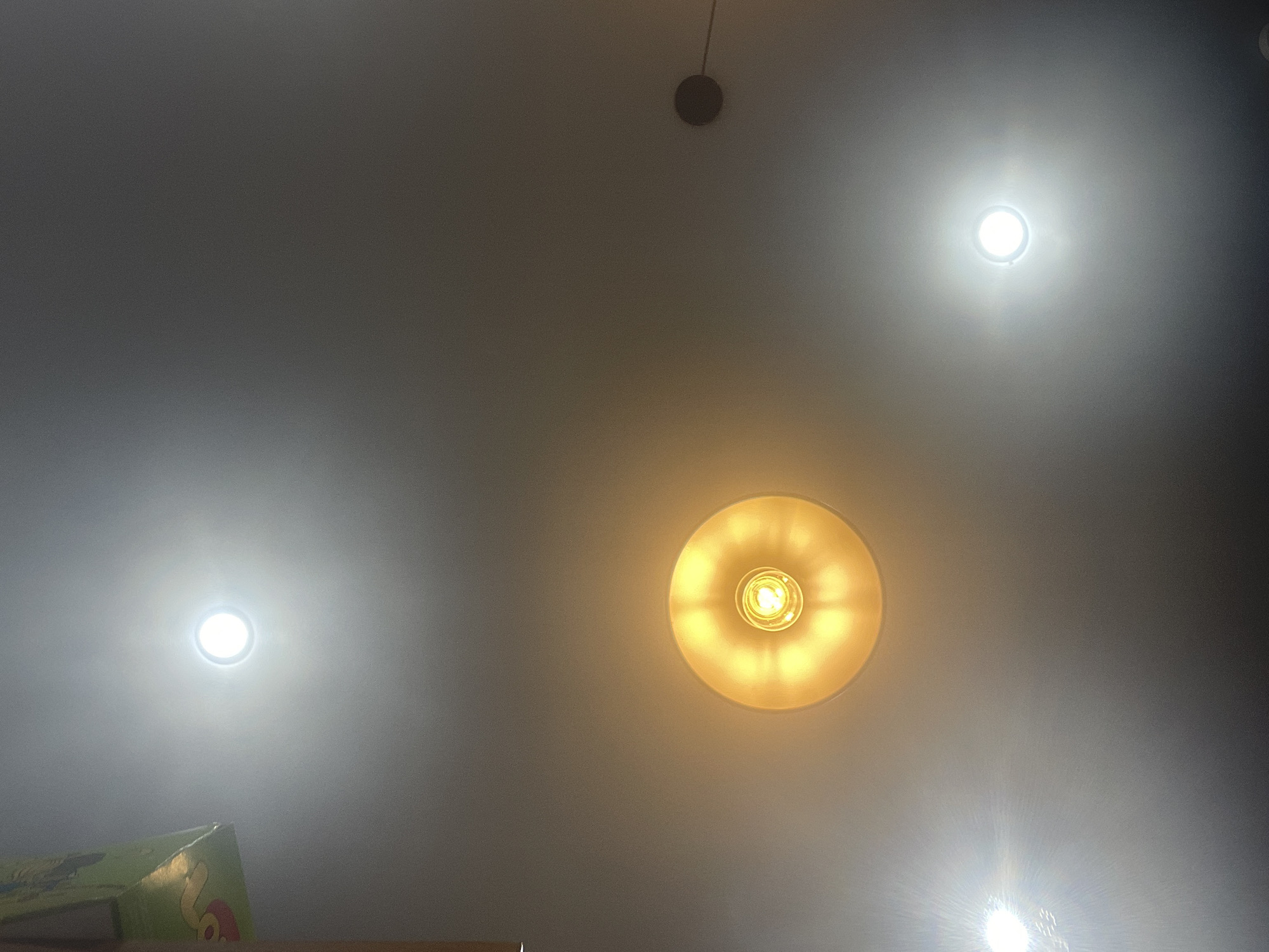 The image shows a ceiling with several lights. In the top left corner, a portion of a green box with yellow lettering is visible. There are three visible light sources on the ceiling: a bright white circular light in the upper middle, a warm yellow circular light with visible filament in the middle, and another bright white circular light in the lower right. A small dark circular object is visible in the far right of the image, attached to the ceiling by a thin wire. The overall scene is indoors, illuminated by artificial light. There are no people or animals present. There is no indication that this image is from Шебунино, Russia.