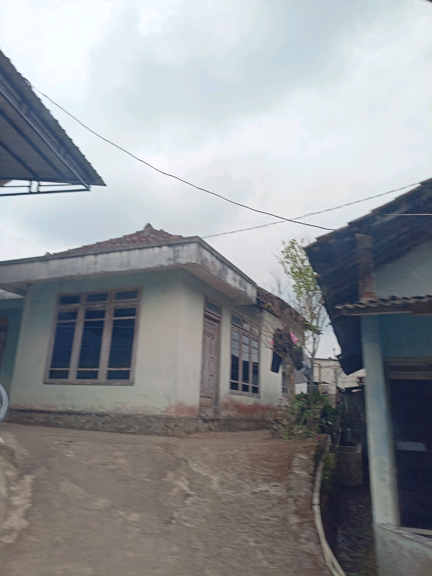 A low-angle, slightly blurred, outdoor, daytime, medium shot captures two houses in Kalegen, Indonesia. The house on the left is pale green with a light brown, tiled roof and a single, wooden door and a large, multi-paned window. The house on the right is partially visible, appearing to be light blue with a dark, tiled roof. A paved pathway slopes upwards towards the left house. A thin, black wire stretches across the top of the frame. The sky is overcast and grey. There are no people visible in the image.