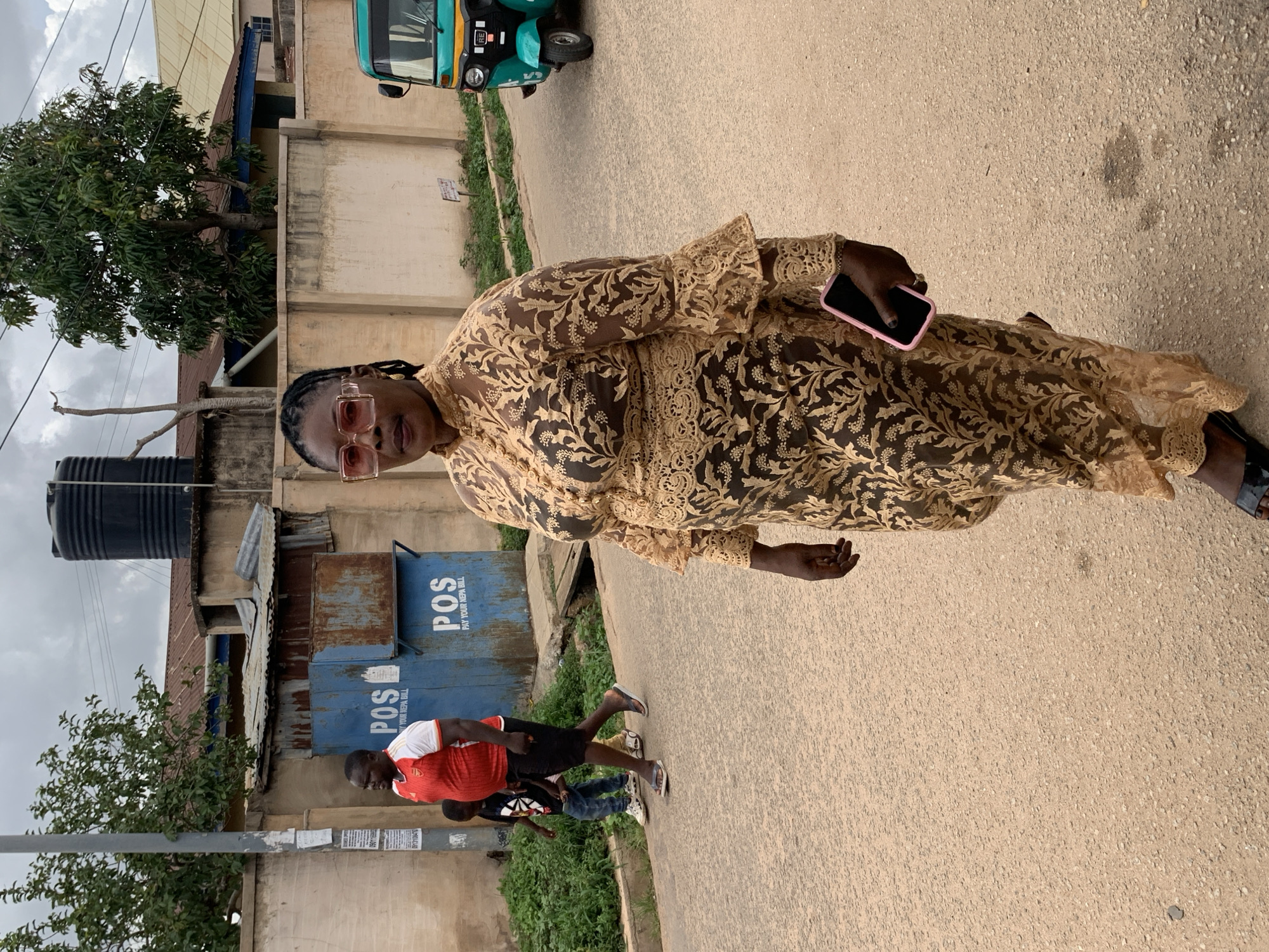 An adult woman in an embroidered gown and glasses is holding a cell phone, standing on a paved road in a suburban area. In the background, several people are walking along the side of the road near metal utility boxes with "POS" printed on them. In the background there is also an auto rickshaw and a building.