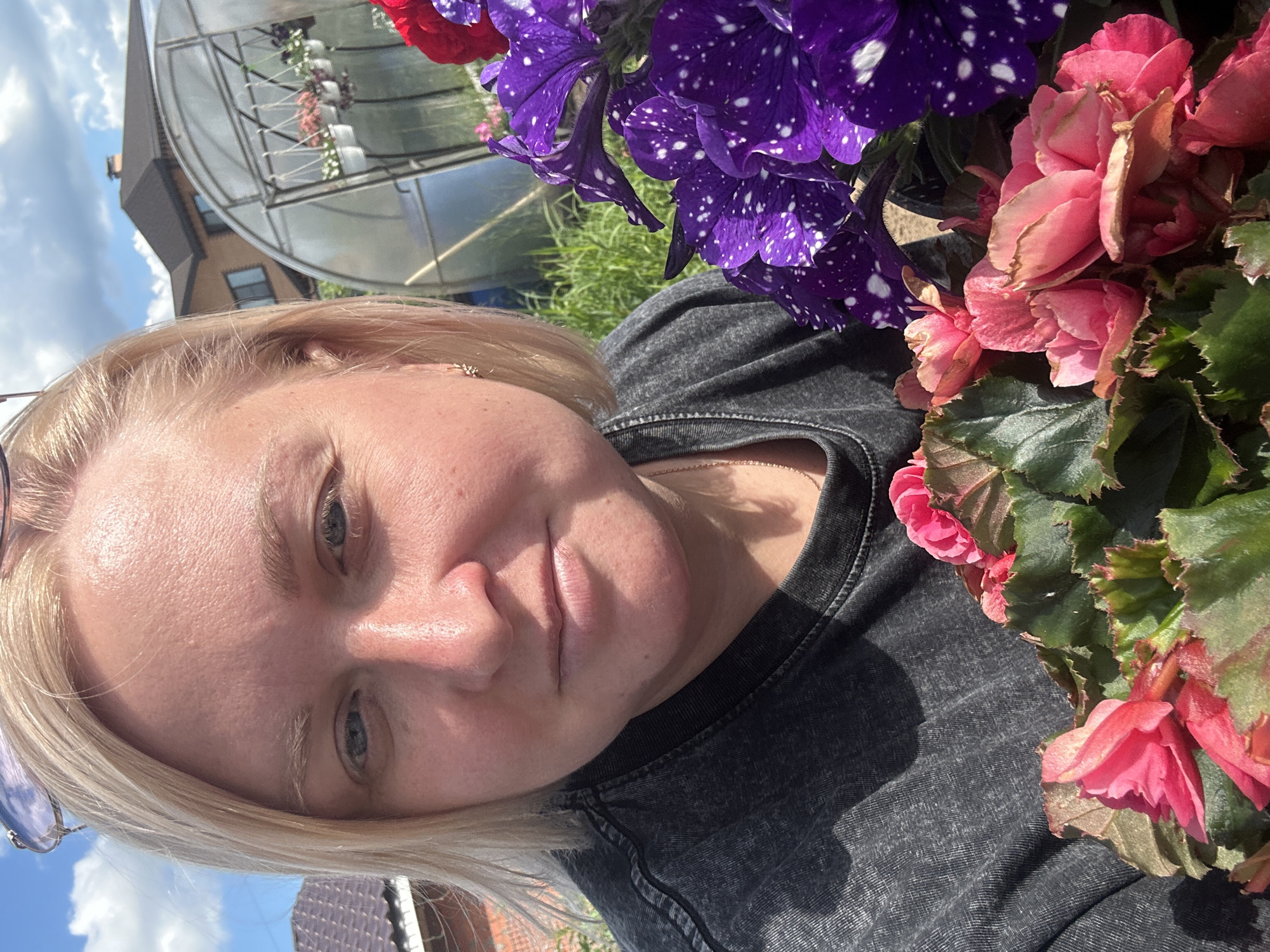 The image shows a close-up, tilted selfie of a blonde woman in an outdoor setting. She is wearing a dark gray t-shirt. In the foreground, to the right of the woman, are several pink begonia flowers and lush green leaves. Behind her, to the left, is a greenhouse structure with hanging plants and a building with windows. The sky is visible with white clouds, suggesting it is daytime and likely sunny. The woman's expression is neutral, and the lighting indicates it is a bright day. The scene appears to be a garden or a place with a greenhouse. The provided location context is Chaikovsky, Russia. There is no visible text in the image.