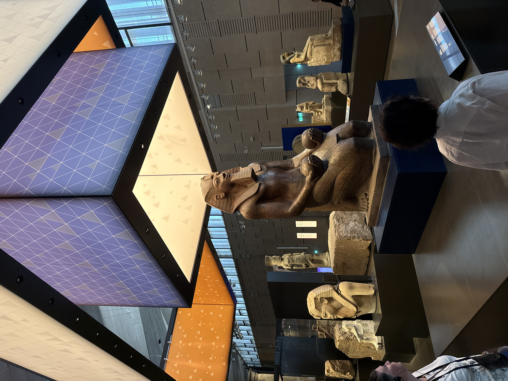 The image shows an interior view of a museum exhibit, likely featuring ancient Egyptian artifacts. In the foreground, two individuals are observing the displays. The person on the left, with dark hair and wearing a white shirt, is partially visible from the side, looking towards the exhibits. The person on the right, also with dark hair, is seen from behind, wearing a white shirt and headphones, and is looking at a digital display.
The central focus of the exhibit is a large, seated statue of an ancient Egyptian figure, likely a pharaoh, made of dark stone. The statue is depicted wearing a traditional headdress and beard, and it holds two spherical objects in its lap. To the left of this main statue, a bust of another Egyptian figure is displayed on a dark pedestal. Scattered throughout the middle ground are other stone sculptures and fragments, some standing upright and others resting on pedestals.
The museum environment is modern, with a high ceiling featuring large, illuminated panels with geometric patterns in orange and purple. The walls in the background are textured and feature vertical lines. The lighting is a mix of ambient light from the ceiling panels and spotlights directed at the exhibits. The floor is dark and reflective. There is no visual indication of the specific location within the city of Kafrat Nassar, Egypt, as the image focuses on the museum's interior.