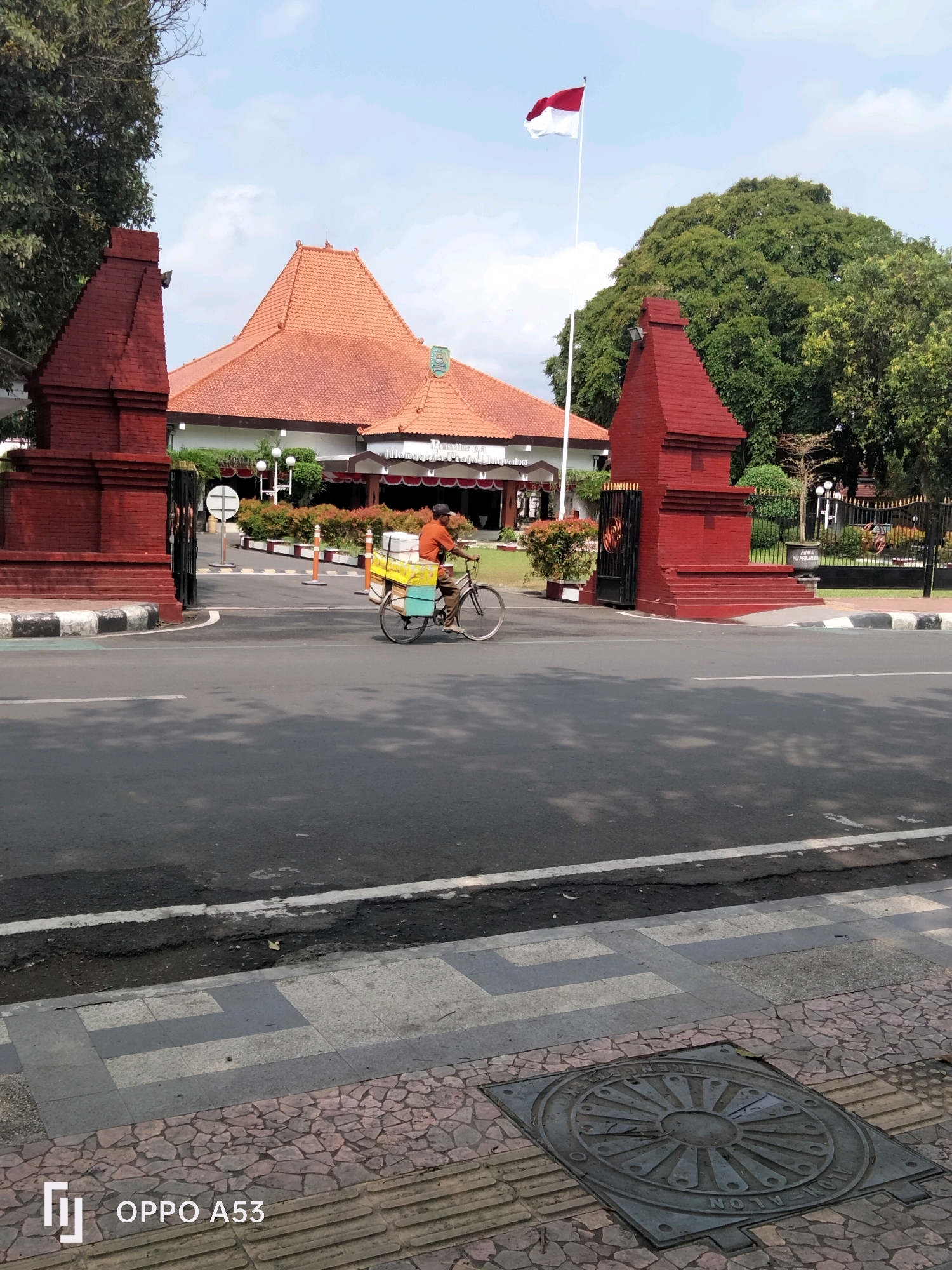 The image shows an outdoor scene in front of a building with red tiled roofs and red brick gate posts. A person on a bicycle, carrying stacked boxes, is moving from left to right in front of the building. The building appears to be a government or cultural institution, with a flagpole flying the Indonesian flag. The street in the foreground is paved, with sidewalks made of patterned tiles and stone. The location is identified as the Trenggalek City Hall in Trenggalek, Indonesia. The weather appears to be clear and sunny.