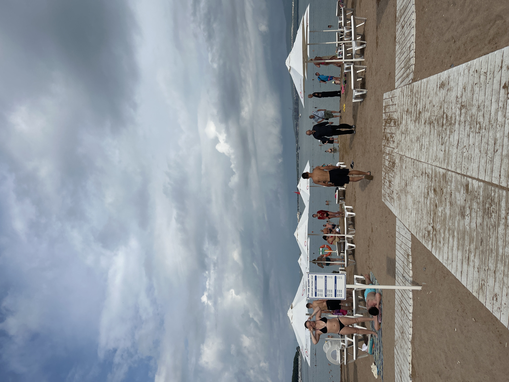 This image depicts a beach scene in Gelendzhik, Russia, on a cloudy day. The beach is sandy with white wooden boardwalks leading to the water. Several white umbrellas are set up on the sand, with lounge chairs underneath.
In the foreground, a person in a black bikini is standing on the left, adjusting their hair. To their right, a person in blue shorts is lying on a towel. A wooden sign with Cyrillic text and a price list is positioned between them. Further down the boardwalk, a shirtless man in black shorts is walking away from the camera towards the water.
In the midground, a group of people are gathered near the water. Some are standing, while others appear to be swimming. One man in a dark shirt and another in a light blue shirt and khaki shorts are walking towards the right. A woman in black and a woman in a blue outfit are also visible. Several white lounge chairs are arranged near the umbrellas.
In the background, the sea stretches out, with a few boats and a crane visible in the distance. The sky is heavily overcast with dark, dramatic clouds. The overall atmosphere is one of a public beach with a moderate number of visitors.