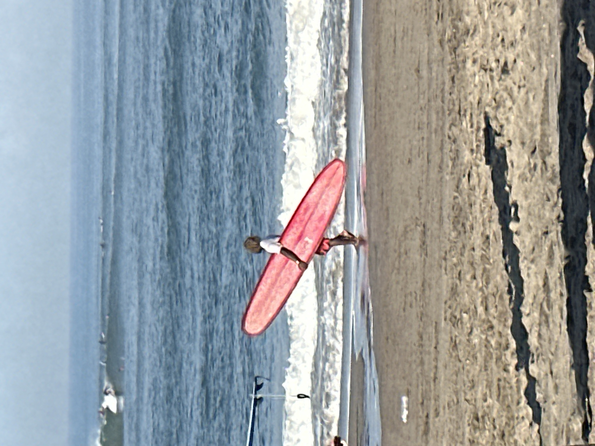 A person with light-colored hair and a white shirt is walking along the sandy shore of a beach, carrying a long, bright pink surfboard under their right arm. The person is walking towards the right side of the frame. The ocean is visible behind them, with small waves breaking and several surfers in the distance. The sky is hazy and blue. The sand in the foreground shows footprints and shadows cast by unseen objects. A partial view of a blue and white object, possibly another surfboard or part of a boat, is visible on the left edge of the frame. The scene appears to be a sunny day at a popular surfing spot. This is likely located in North Kuta, Indonesia, a region known for its beaches and surf culture.