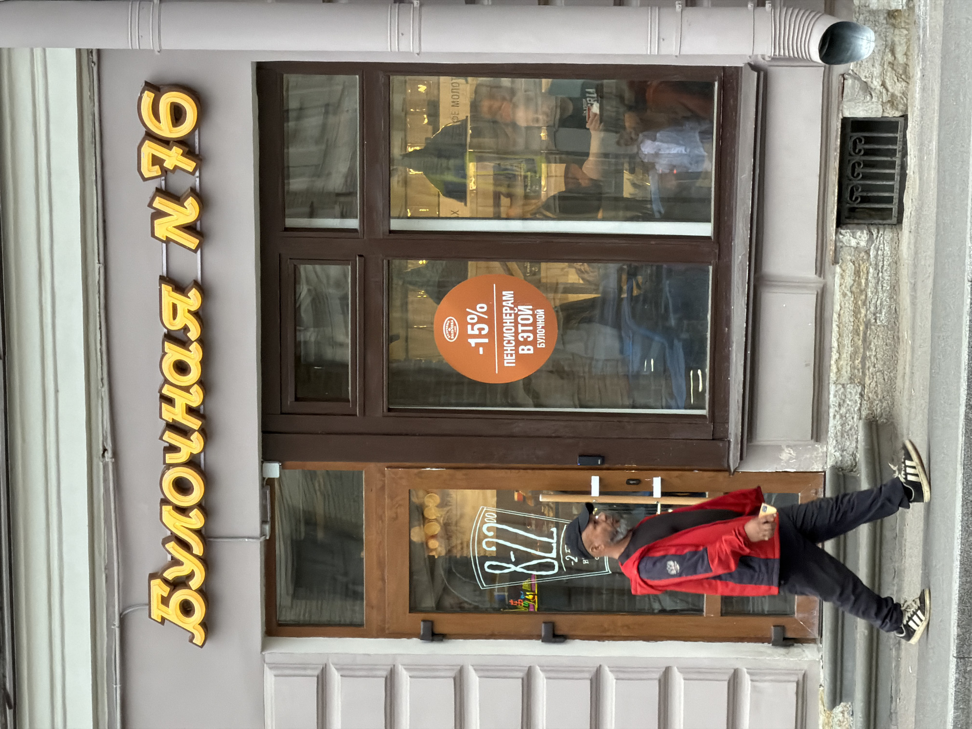 Here is a description of the image:
The image captures a street scene in Saint Petersburg, Russia, in front of a bakery (булочная). The bakery's sign, "Булочная N 76," is displayed vertically in bright yellow letters on the left side of the image. A man wearing a red jacket, black pants, and black-and-white sneakers is walking past the bakery. He is holding something in his right hand.
The bakery has a large window divided into six panes. A brown sticker advertising a 15% discount for pensioners is visible on the lower pane. Reflections in the window show activity inside the bakery. The entrance door, also wooden, is visible next to the window, with a graphic showing the opening hours "8-22" emblazoned on it.
Above the window, a downspout runs vertically along the wall. To the right of the window is a small, grated opening in the building's exterior wall. The overall setting suggests a typical urban street scene in Saint Petersburg.