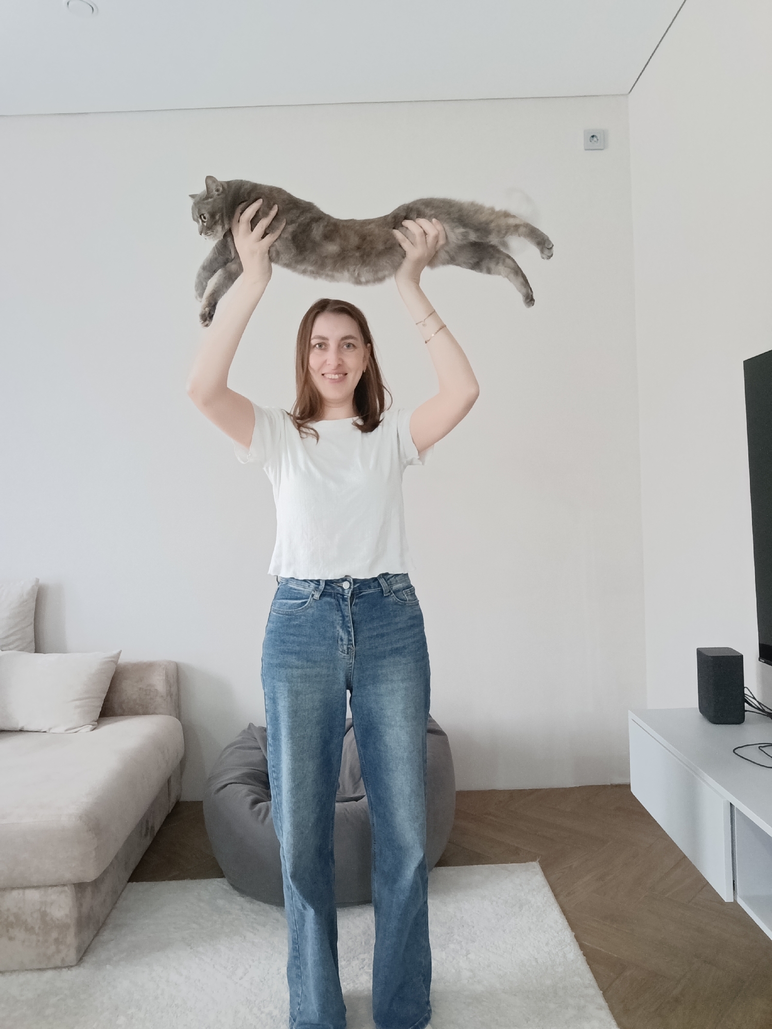 A woman stands in a room, holding a gray cat above her head with both hands. She is wearing a white t-shirt and blue jeans. The room has a white wall behind her, a beige sofa to the left, a gray beanbag chair near her feet, and a white TV stand with a black speaker to the right. The location is Chaikovsky, Russia.