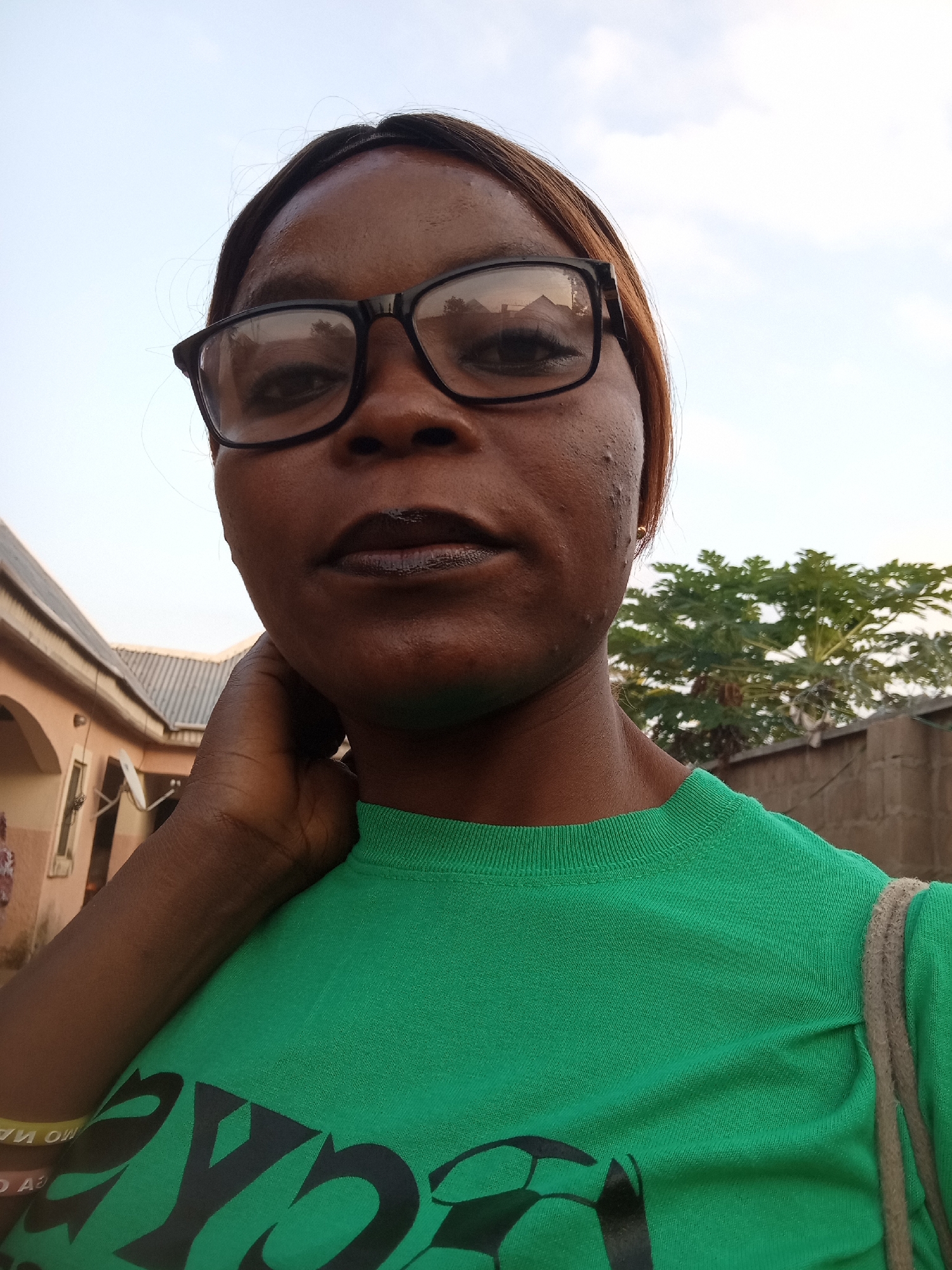 A young woman with dark skin, wearing black-rimmed glasses and a green t-shirt, is pictured from the chest up. She is looking directly at the camera. Her right hand is raised to her neck, and her left arm is down. The background shows a building with arched entrances on the left, and a brick wall with greenery behind it on the right. The sky is overcast. The location is Kason, Nigeria.