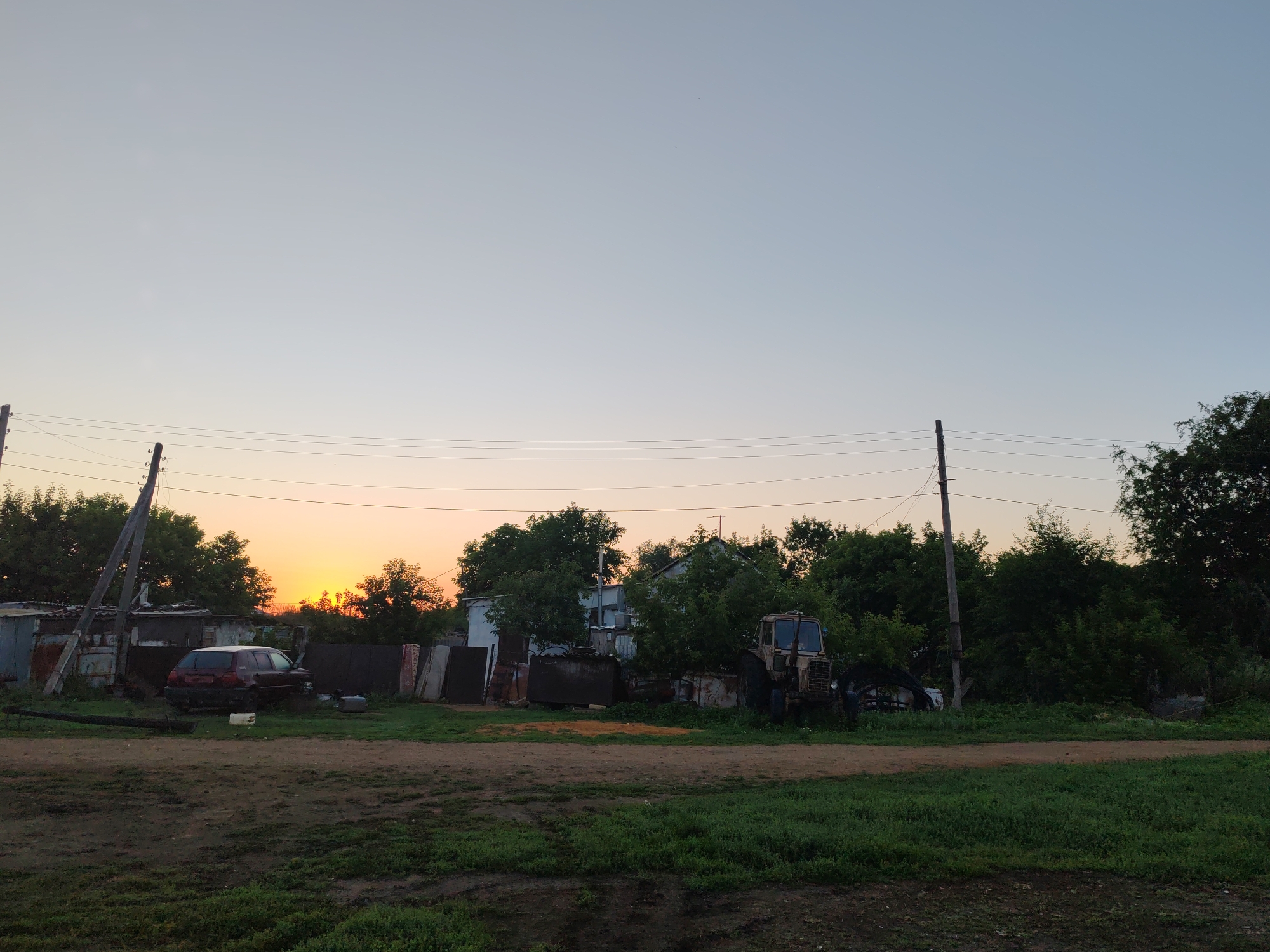 A wide-angle shot captures a rural scene in the city of Akzhar, Kazakhstan, at dusk. The sky is a pale, gradient blue transitioning to a soft orange glow near the horizon, indicating the setting sun. Several power lines stretch across the upper half of the frame, supported by wooden poles. In the middle ground, a collection of structures and vehicles are visible. To the left, a dark red hatchback car is parked near some makeshift sheds and fencing. Further to the right, a weathered, light-colored tractor sits amongst dense green foliage and more scattered outbuildings. The foreground consists of a dirt path and a grassy verge. The overall impression is one of a quiet, somewhat overgrown, and utilitarian rural settlement under a fading sky. There are no people or animals visible in the frame.