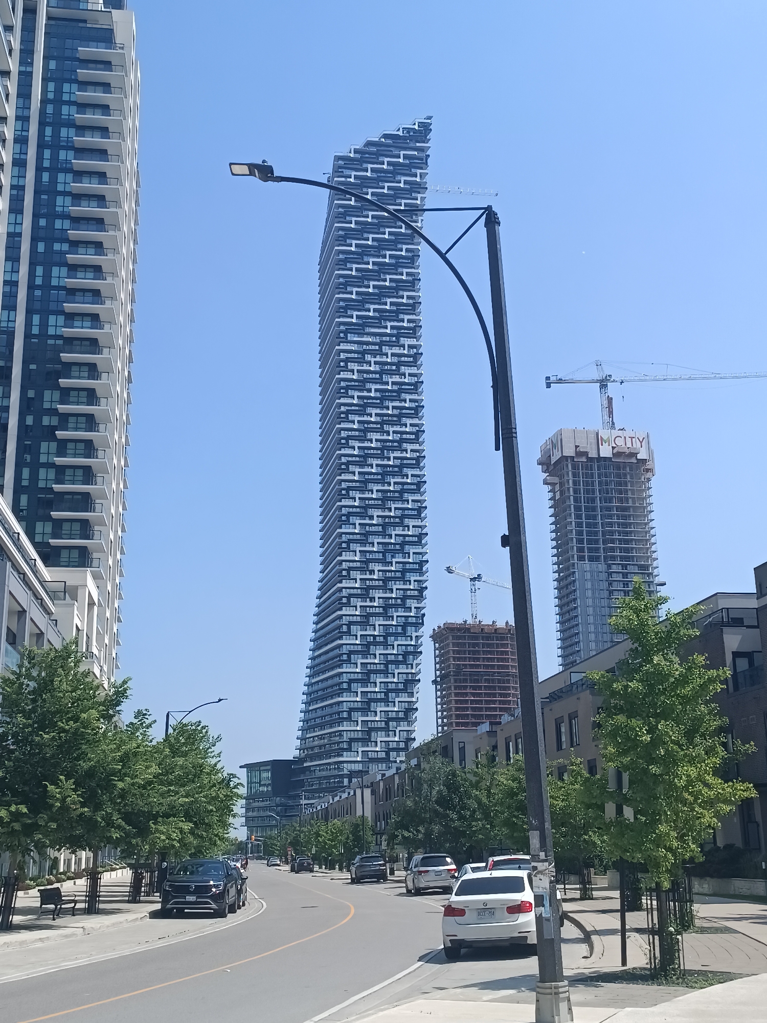 The image depicts a street scene in Mississauga, Canada, likely in the Square One District, an area known for its ongoing development of high-rise condominiums. In the foreground, a curved road leads into the distance, bordered by sidewalks and mature trees. Several cars are visible on the road, including a dark SUV parked on the left and a white sedan in the right lane. Along the left side of the street, a row of modern townhouses or low-rise condominiums is visible, with benches and street lamps lining the sidewalk.
Dominating the background are several tall buildings. To the far left, a completed residential skyscraper with a white facade and numerous balconies is prominently featured. To its right, a distinctive, wavy skyscraper with a modern, layered design is a central focus. Further in the background, a skyscraper under construction, labeled "M.CITY," is visible with a construction crane overhead, indicating ongoing development. Another, shorter building under construction is also present. The sky is clear and blue, suggesting a sunny day. The overall impression is of a rapidly developing urban environment.