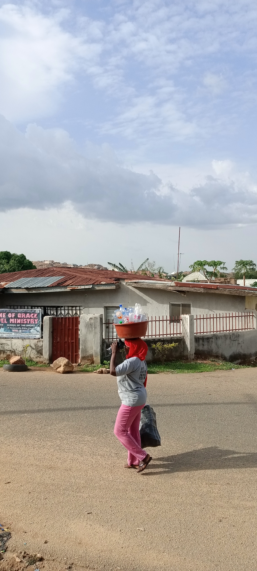 A woman is walking on a street in the city of Unknown, Unknown, carrying a red basin full of plastic water bottles on her head. She wears a red headscarf, gray shirt, and pink pants, and is carrying a black bag in her left hand. The street is a paved road with a dirt shoulder. Behind her is a building with a rusty red roof. The building has a sign that reads "NE OF GRACE" and "PEL MINISTRY". There are trees and other buildings visible in the background. The sky is cloudy.