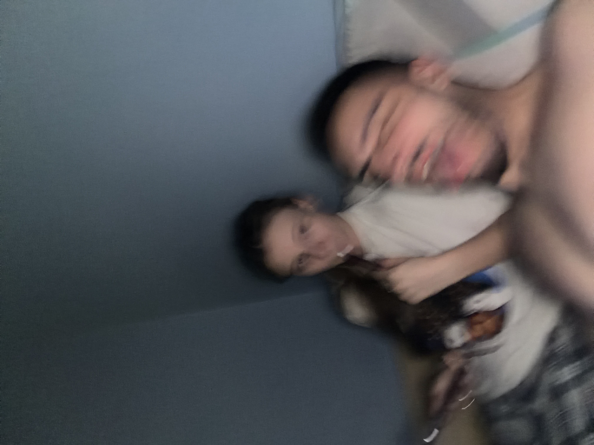 The image is blurry and appears to depict two people indoors, potentially in a bedroom setting. A person with short dark hair is in the foreground, face distorted and mouth open. A second person is in the background, wearing a light-colored shirt. A hand is visible near the second person. The background includes a blue wall and white bedding. The location cannot be determined from the image.