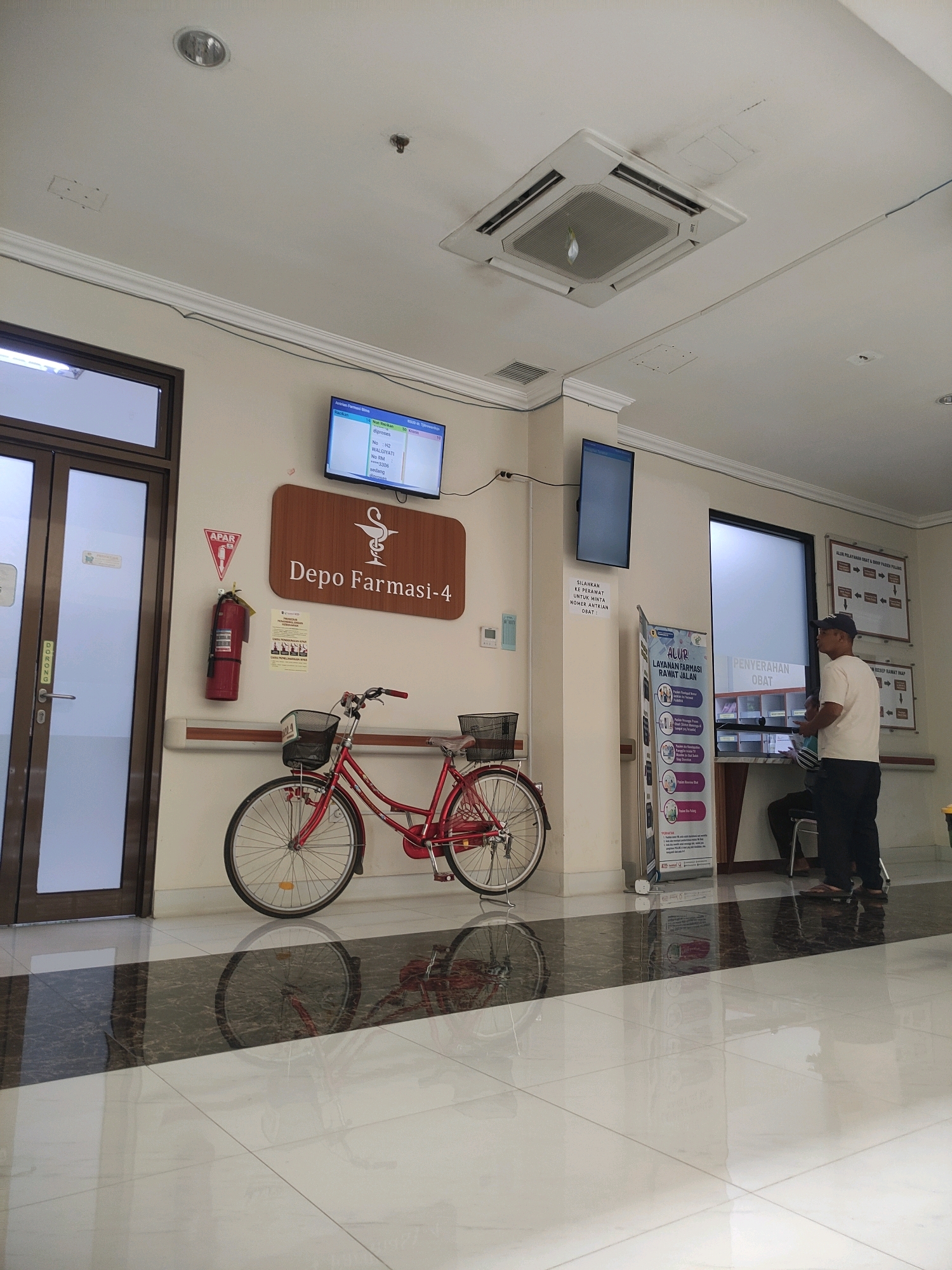 The image depicts the interior of what appears to be a pharmacy or medical facility in Purworejo, Indonesia. In the foreground, a red bicycle with two baskets is parked on a polished floor with a reflective surface. To the left, a glass-paneled door with "DORONG" (push) written vertically is visible. On the wall to the right of the door, there's a fire extinguisher, a sign with a red triangle and "APAR" (fire extinguisher), and a wooden sign with a Caduceus symbol and "Depo Farmasi-4" (Pharmacy Depot-4). Above the wooden sign, a flat-screen TV displays queue information. Further to the right, another flat-screen TV is mounted on the wall. A person wearing a dark cap and a light-colored t-shirt stands near a counter, interacting with someone seated. Various informational posters and signs are displayed on the walls, including one titled "ALUR LAYANAN FARMASI RAWAT JALAN" (Outpatient Pharmacy Service Flow) and another that reads "PENYERAHAN OBAT" (Drug Handover). The environment appears to be well-lit with overhead fluorescent lighting and a visible air conditioning unit in the ceiling. The floor is tiled with a black and white pattern that creates a strong reflection of the objects and people present.
