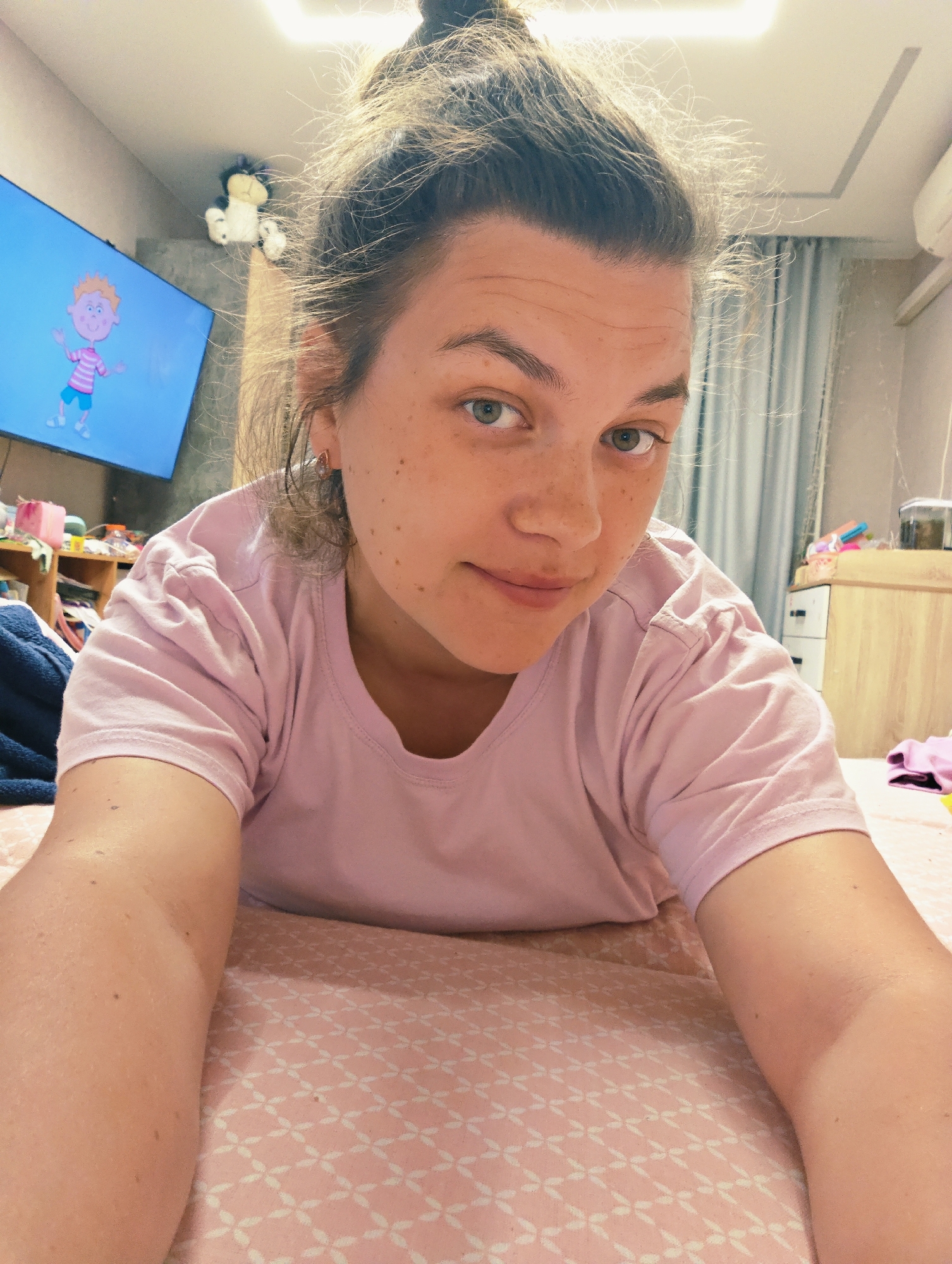 A young adult female is depicted in a close-up, low-angle selfie, lying prone on a bed with a pink and white geometric patterned duvet. She has fair skin with numerous small freckles visible on her nose, cheeks, and forehead. Her light brown hair is pulled up into a messy bun, showing loose strands. Her eyes are light green/hazel and gaze directly at the camera, while her mouth is closed with a subtle, soft smile. She wears a plain, light pink short-sleeved t-shirt and a small, light-colored earring in her visible right earlobe.
The background reveals an indoor bedroom setting, located in Chaikovsky, Russia. To the upper left, a wall-mounted flat-screen television displays a brightly colored animated male character with orange hair, a pink and white striped shirt, and blue shorts. Above the television, a black and white plush toy, possibly a cat or dog, is perched on a white shelf or ledge near the ceiling. Further left, a wooden shelving unit holds various indistinct items and a dark blue blanket.
On the right side of the frame, light blue/grey pleated curtains are visible. A white wall-mounted air conditioning unit is affixed to the wall near the ceiling. Below it, a light wooden dresser with white drawers is present, topped with a small transparent container (possibly an aquarium or jar) and some folded pink clothing. The white ceiling features a rectangular recessed design and a prominent linear light fixture providing bright illumination to the scene.