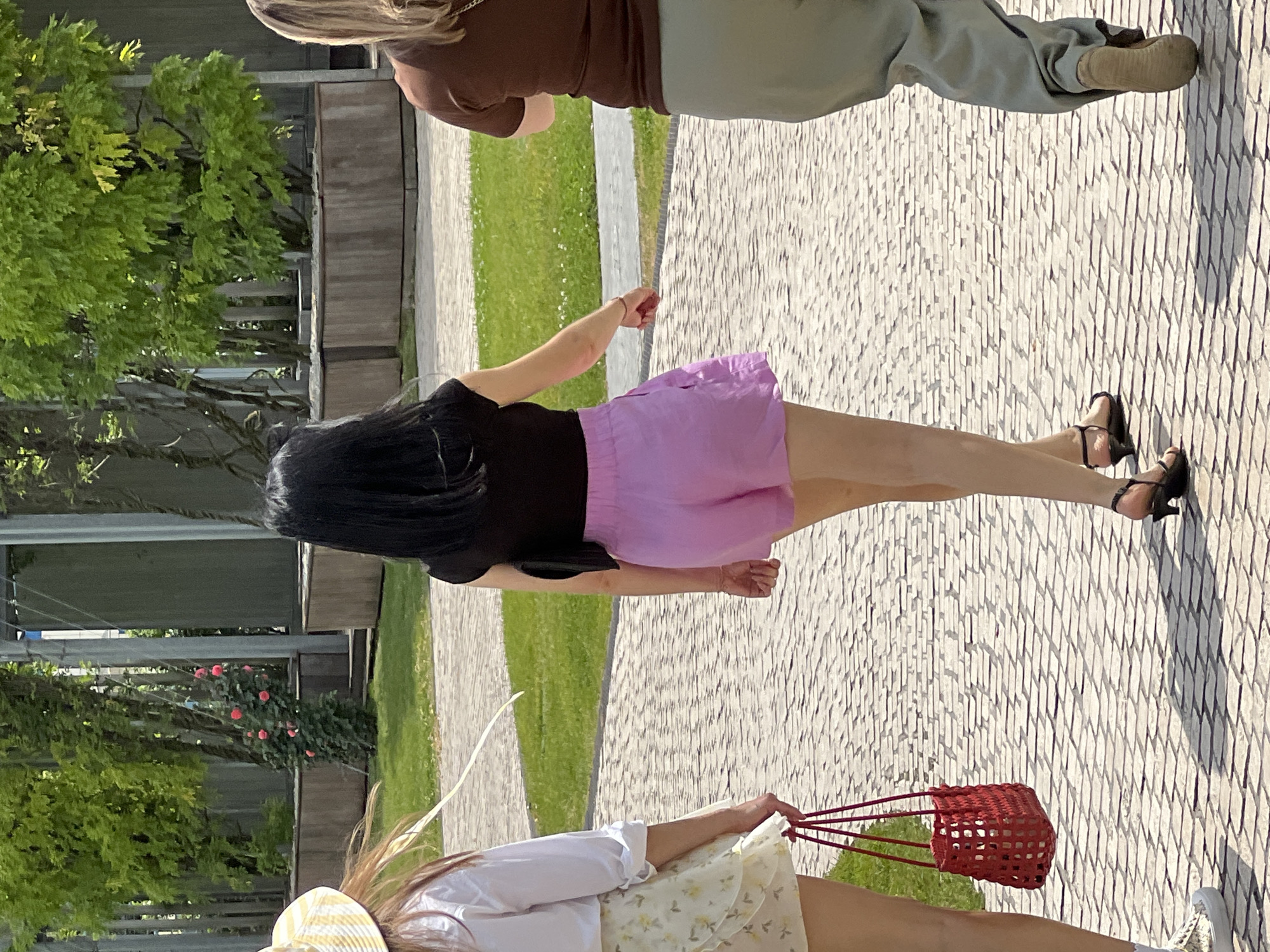 Three individuals are walking away from the camera on a paved walkway. The person in the center has long black hair and is wearing a black t-shirt and pink shorts. The person on the left has long brown hair and is wearing a white shirt and a light-colored floral skirt, carrying a red woven bag. The person on the right has blonde hair and is wearing a brown t-shirt and light green pants. The walkway is lined with grass and trees in the background, and there are planter boxes. The scene appears to be an outdoor public space, possibly a park or garden, illuminated by bright daylight. There is no visible indication of the location being Krasnodar, Russia, or any other specific city.