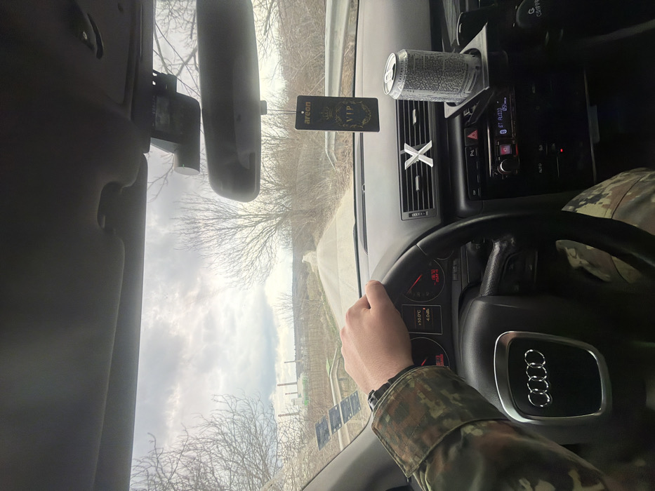 The image depicts the interior of an Audi vehicle from a front-seat perspective, looking forward through the windshield onto a rural road near Opanets, Bulgaria. The road, likely unpaved or gravel, recedes into the distance, flanked by bare deciduous trees, suggesting a non-summer season. In the far background, an industrial facility with multiple chimneys, consistent with the Opanets Thermal Power Plant, is visible under a cloudy sky.
The driver's right hand, clad in camouflage-patterned attire and wearing a dark-colored watch, rests on the top of the Audi steering wheel. The vehicle's dashboard shows an external temperature reading of +10.0°C. An aftermarket car stereo displays "BT Audio" and "87.50". Hanging from the rearview mirror is an "Areon VIP" car air freshener. A dashcam is mounted behind the rearview mirror. A silver "X" ornament is affixed to the central air vent on the passenger side. An aluminum beverage can, predominantly white and red with Cyrillic text, is secured in a black aftermarket cup holder attached to the central console. Below the cup holder, a "GRIPPER" branded phone mount is visible.