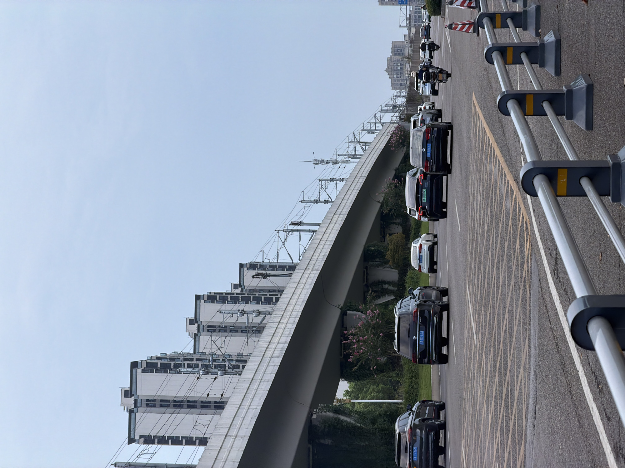 This is a vertical, eye-level, outdoor shot of a multi-lane road with traffic in Jiangjin, China. The sky is overcast and pale blue. On the left side of the image, an elevated structure, possibly a highway or railway overpass, runs horizontally across the frame. The overpass is supported by concrete pillars, some of which are covered in ivy. A line of modern, multi-story residential buildings with white facades and dark windows is visible behind the overpass.
The road itself has multiple lanes marked with yellow and white lines. Several cars are driving on the road, including a black SUV on the left, a black Volkswagen SUV in the middle, and a white car on the right. Further down the road, more cars and a motorcycle with a rider wearing a helmet are visible. Traffic cones and a metal guardrail are present in the foreground. The overall impression is one of a busy urban environment.