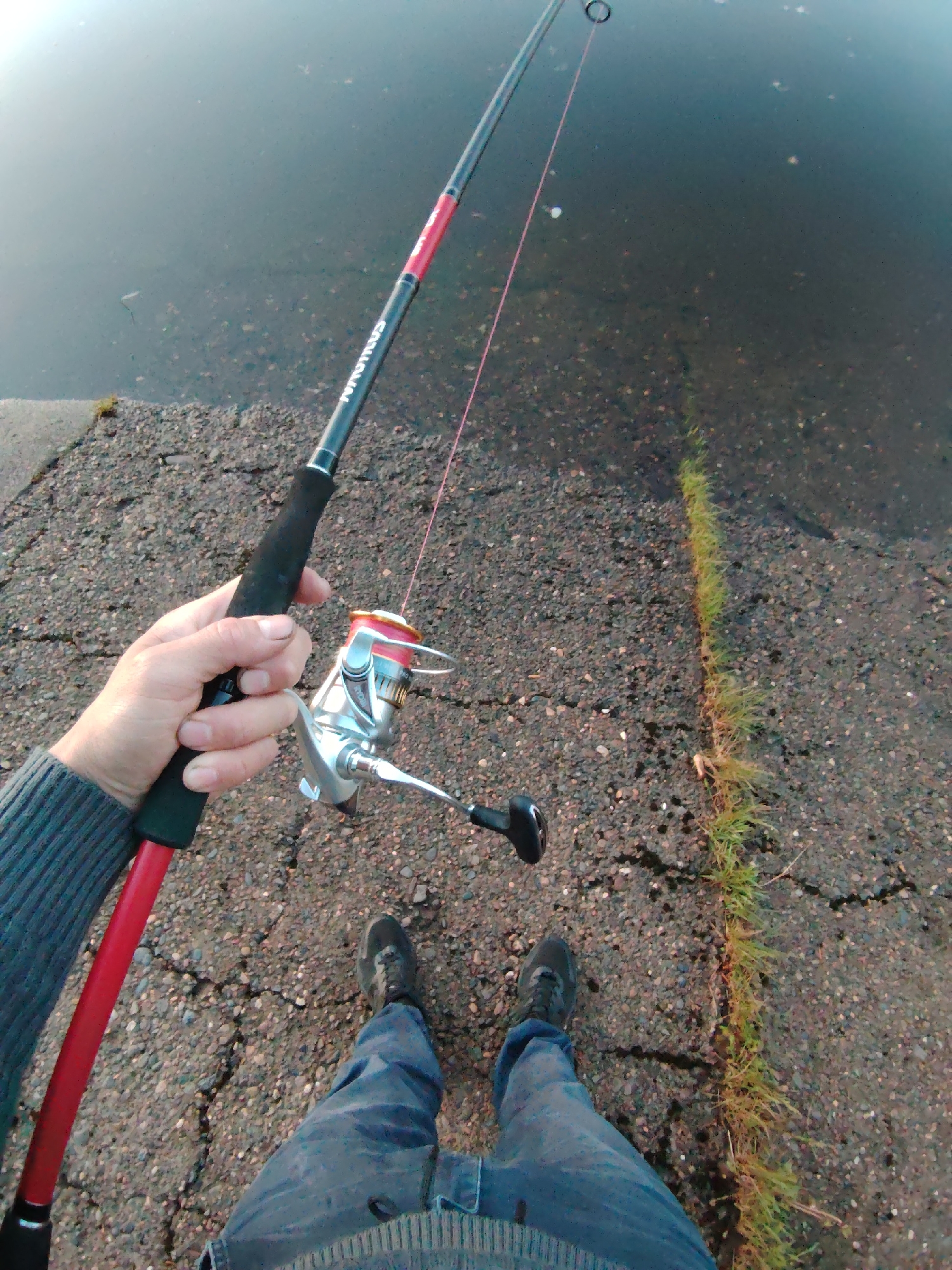 A person stands near a body of water in Chaikovsky, Russia, holding a red and black fishing rod with a silver reel. The person wears blue jeans, a gray sweater, and black shoes. The ground is cracked and covered with small pebbles. A line of grass grows in a crack in the ground. The water appears dark.