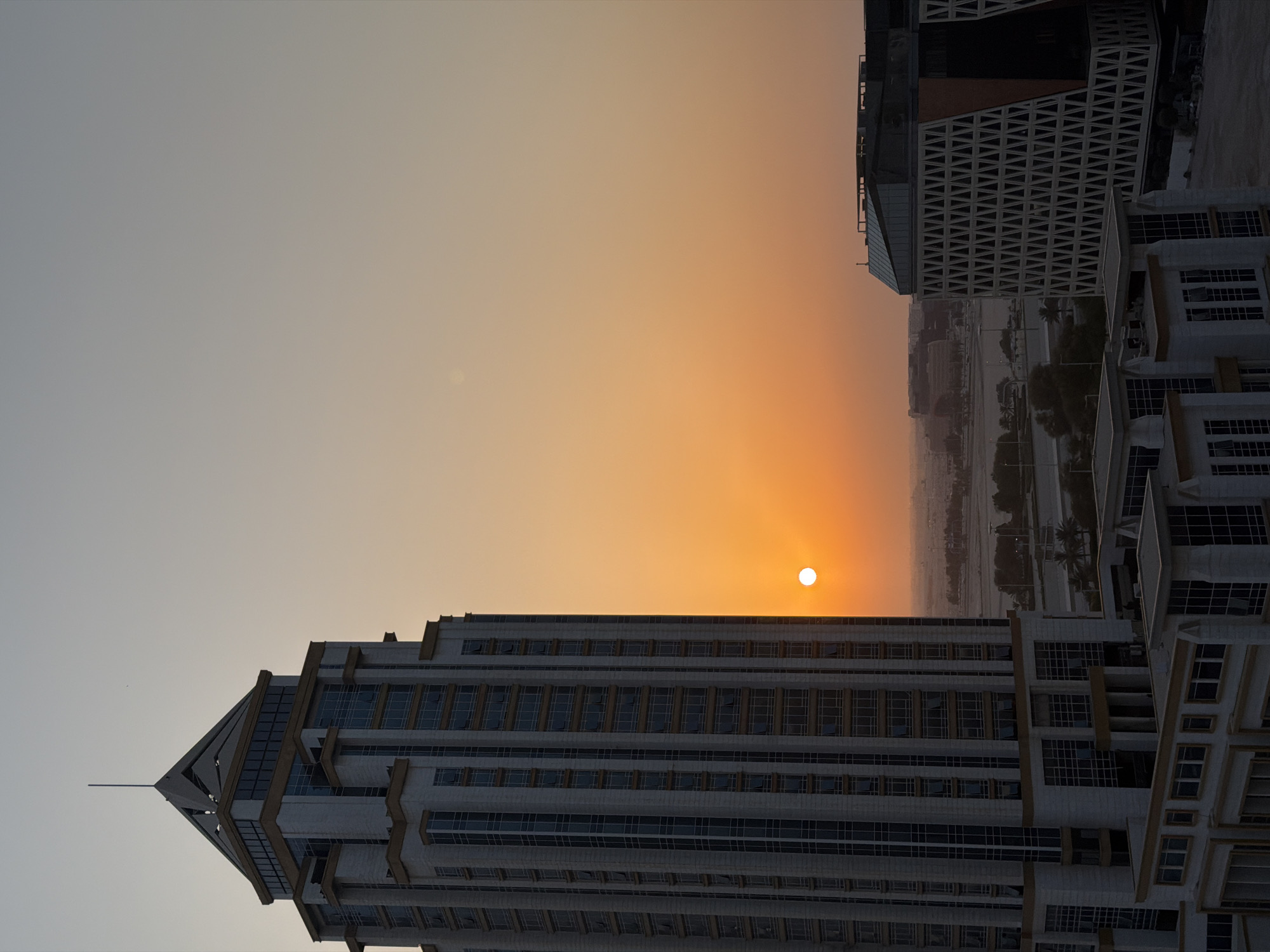 This image captures a panoramic view of the cityscape of Lusail, Qatar, during sunset. The sun, a bright orange orb, is positioned in the center of the frame, casting a warm glow across the sky which transitions from a pale yellow near the horizon to a soft grey at the top. Dominating the left side of the image is a tall, modern skyscraper with a distinctive pyramid-shaped top adorned with a slender spire. The building's facade is a blend of white and grey stone with rows of glass windows. In the foreground and to the right of the skyscraper, another contemporary building with a unique geometric facade, characterized by a pattern of interlocking triangles, is visible. The background reveals a sprawling urban landscape with various structures and what appears to be a waterfront area dotted with palm trees and streetlights. The overall scene depicts a serene urban environment at dusk, with the interplay of light and shadow highlighting the architectural elements of Lusail.