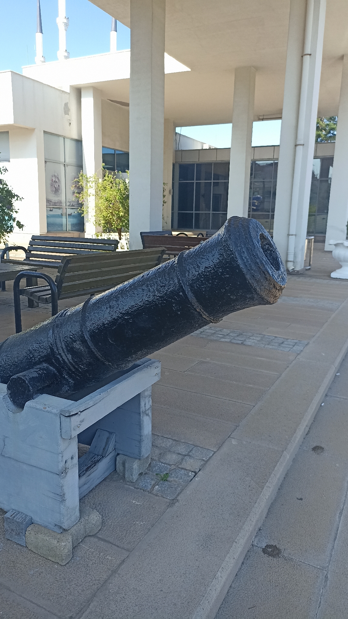 The image shows a black, antique cannon positioned on a light blue wooden stand. The cannon is the primary subject, with its barrel extending diagonally from the lower left to the upper right of the frame. The barrel has a textured, glossy finish and appears to be made of metal. It is mounted on a wooden carriage that is supported by concrete blocks.
In the background, there is a modern building with large windows and white columns. The architecture suggests an outdoor public space. Several wooden benches are visible in front of the building, implying a seating area for visitors. Trees and greenery are present between the benches and the building. The sky is clear and blue, indicating a bright, sunny day. The paving stones in the foreground and midground suggest an outdoor plaza or walkway. The overall setting appears to be a historical site or a public park in a city, possibly near the coast given the imagery seen through the windows, depicting ships. There are no people visible in the frame, and no text is discernible. The time of day appears to be midday, given the bright sunlight and the shadows.