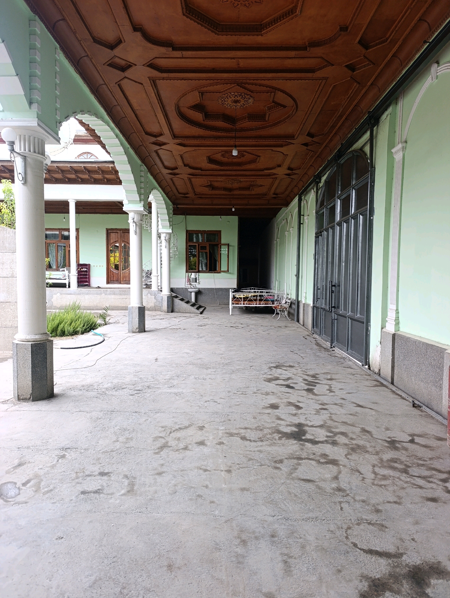 The image depicts a covered walkway with a concrete floor, likely in a residential or historical setting in Margilan City, Uzbekistan. The walkway is lined with pale green walls and supported by white columns with grey granite bases. The ceiling is elaborately decorated with dark wood paneling featuring geometric patterns and ornate carvings. On the left side of the walkway, there is a small garden with green bushes. In the background, a traditional wooden door is visible, along with windows and a white metal bed frame. On the right side, a large set of grey double doors with multiple glass panes dominates the wall. The concrete floor appears damp in places, with dark splotches suggesting water or stains. The scene conveys a sense of traditional architecture and outdoor living space.
