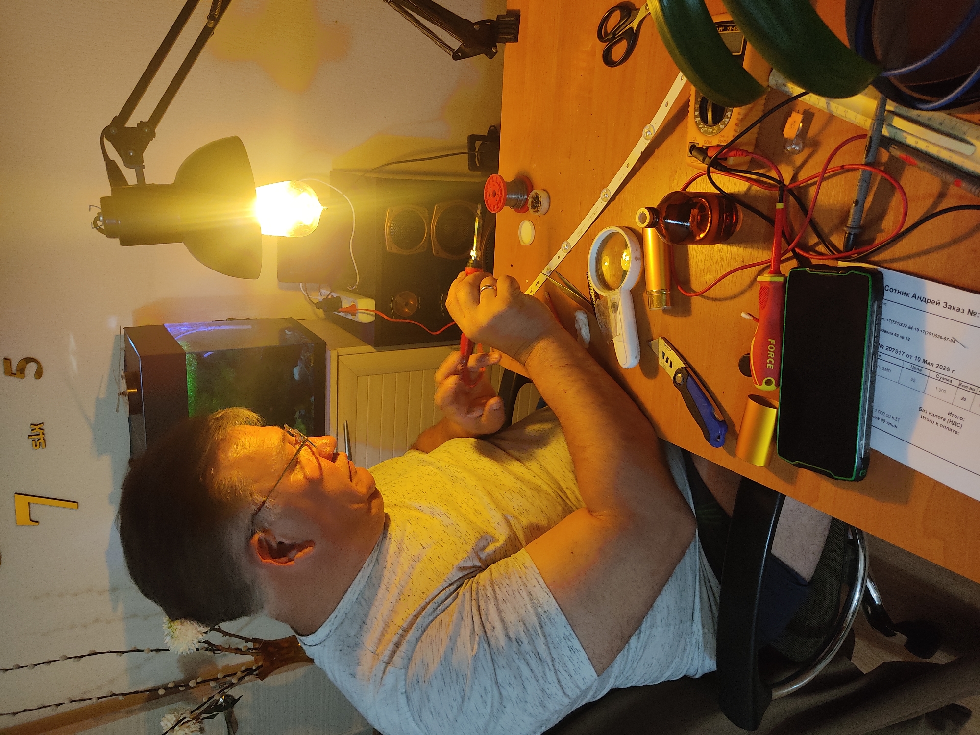 A man is seated at a desk, working on electronics. He is holding a soldering iron and pliers. The desk is cluttered with tools and components, including a magnifying glass, multimeter, and an LED strip. A lamp is positioned above the desk, illuminating the work area. There is a document with Russian text on the desk next to a smartphone. The document appears to be an invoice and includes the name "Сотник Андрей" and a date of "10 Мая 2026 г." A fish tank, speakers, and wall decorations are visible in the background.