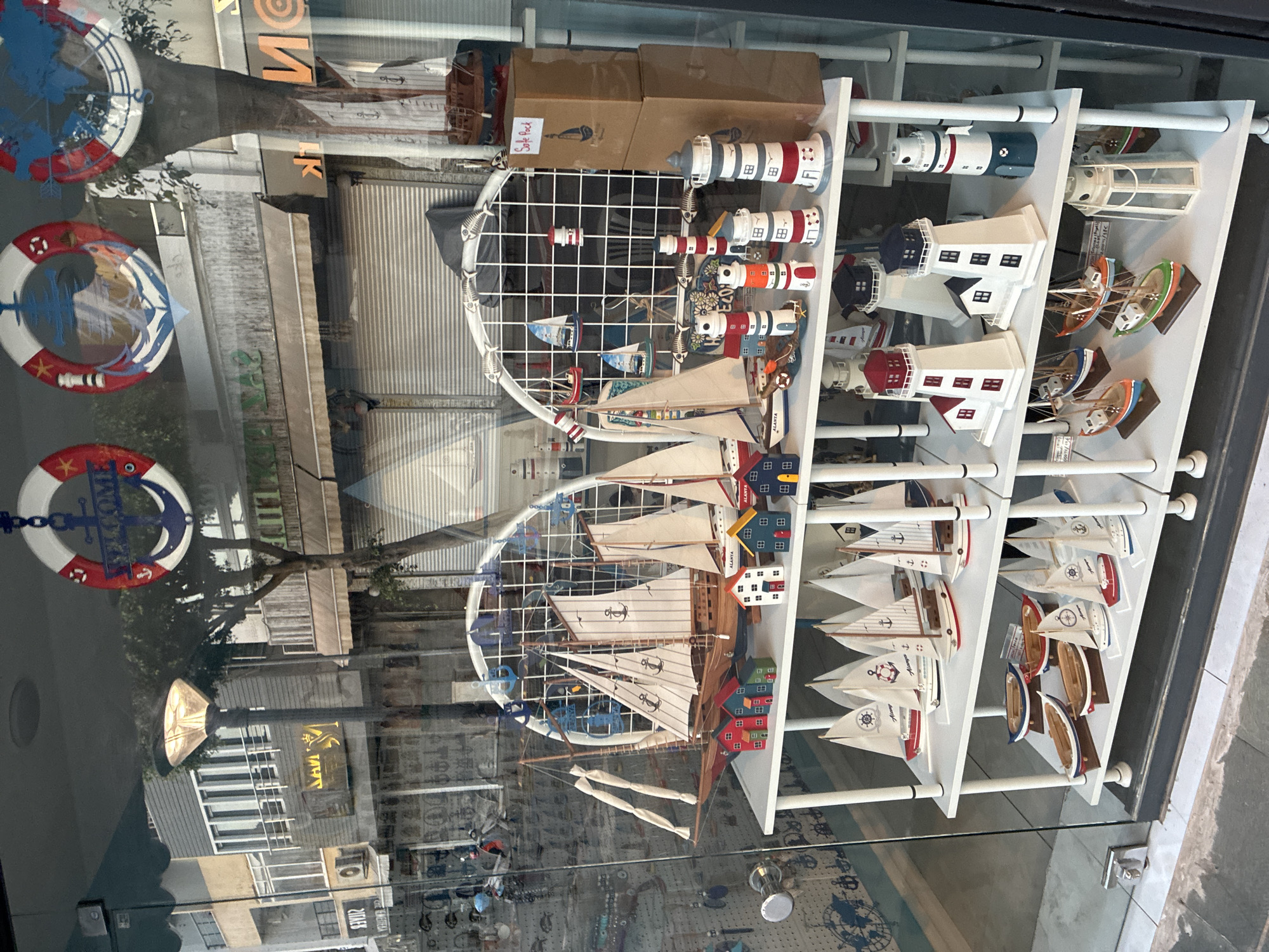 The image shows a store window in Alanya, Turkey, displaying a collection of nautical-themed decorative items. The items include several model sailboats of various sizes, miniature lighthouses in white and red or blue and white, and small wooden houses painted in bright colors. Some of the sailboats are placed on shelves with other miniature boats and decorative elements like anchors and fish motifs. The store appears to be on a street with other buildings visible through the window, including shops with rolled-down metal shutters and awnings. There are also decorative life preservers hanging from the ceiling above the window display. The lighting suggests it is daytime, possibly late afternoon or early morning.