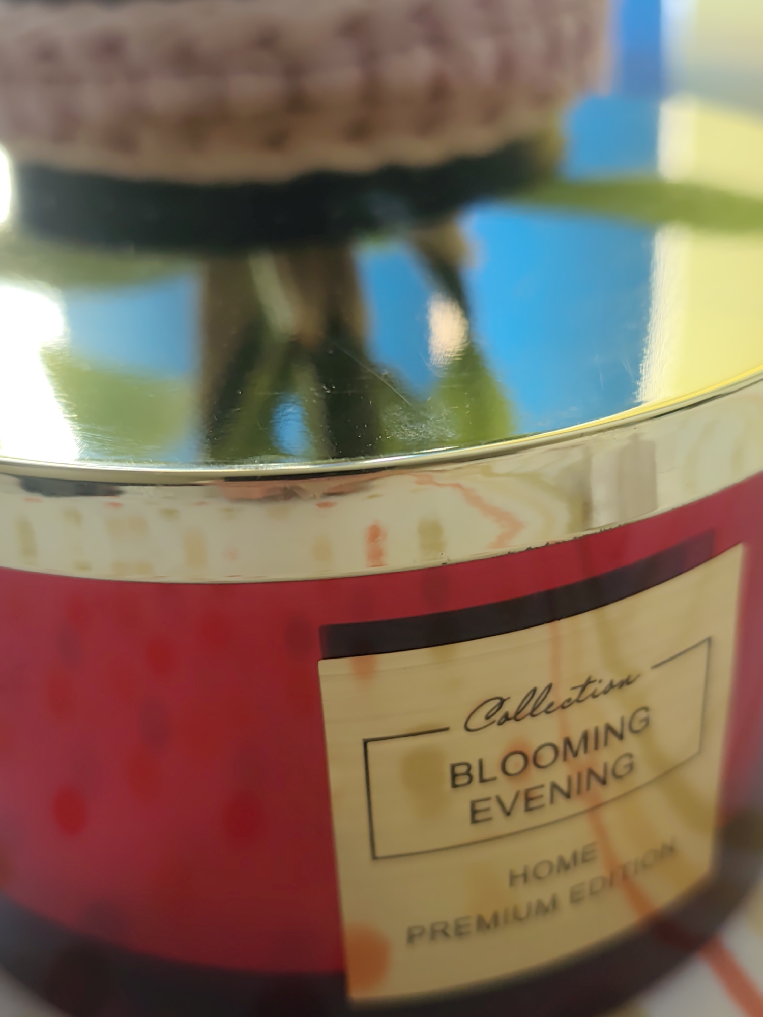 The image shows a close-up of a candle with a red jar and a gold lid. The candle has a label that says "Collection BLOOMING EVENING HOME PREMIUM EDITION." The lid is reflective and shows a blurred background of what appears to be greenery and blue sky, suggesting it might be outdoors or near a window. The lighting is bright, indicating daytime. There are some light reflections on the lid, and the red jar has subtle, lighter red polka dots. The focus is on the candle itself, with the background out of focus.