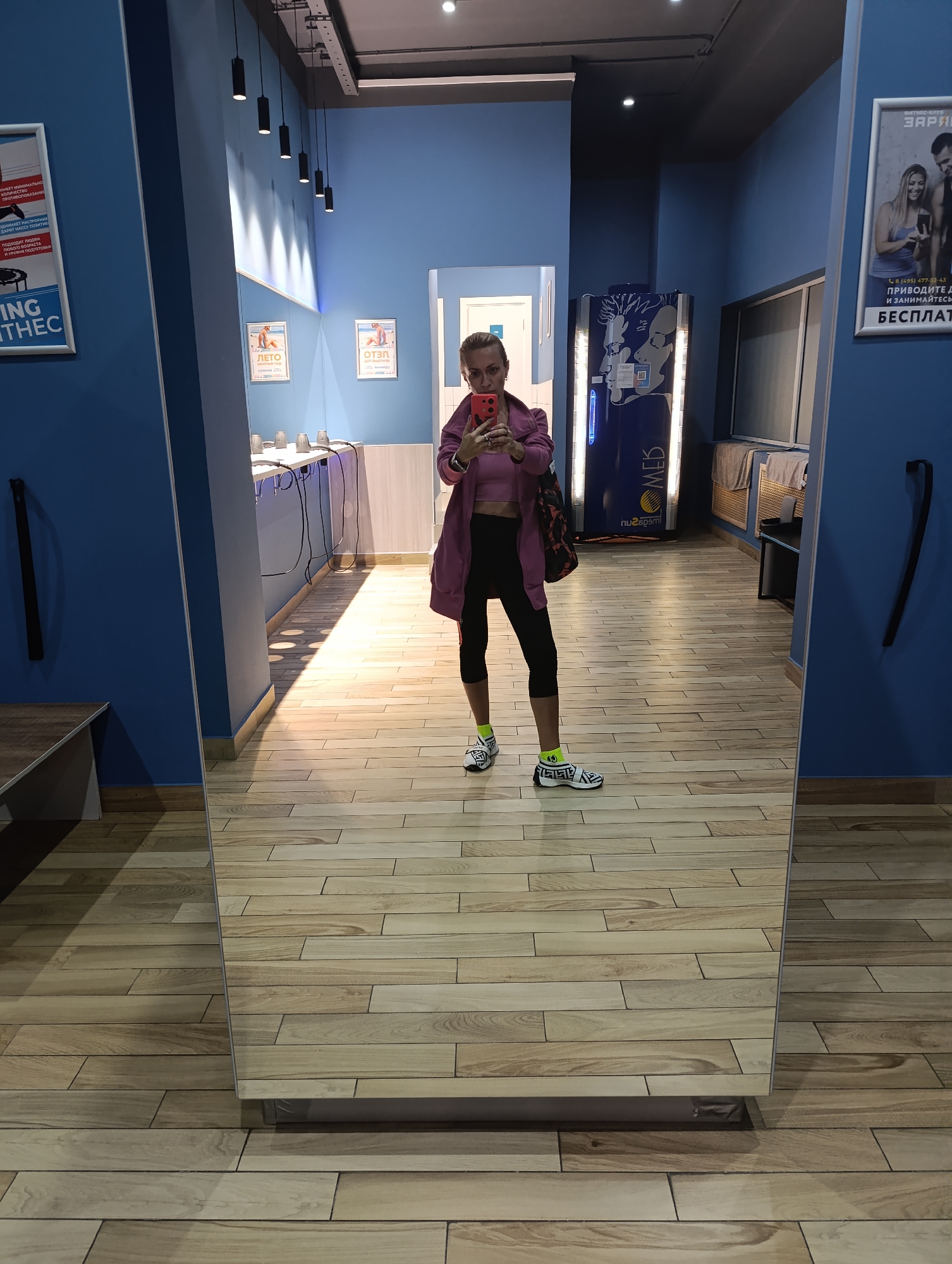 A woman is taking a mirror selfie in a gym locker room. She is wearing a pink crop top, a purple jacket, black leggings, and white and black sneakers with yellow socks. The locker room has blue walls and a wooden floor. There are lockers, a tanning bed, and a vending machine visible in the background. The image is taken from a slightly low angle, and the woman's face is partially obscured by her phone. There is no indication that this scene is in Moscow, Russia.