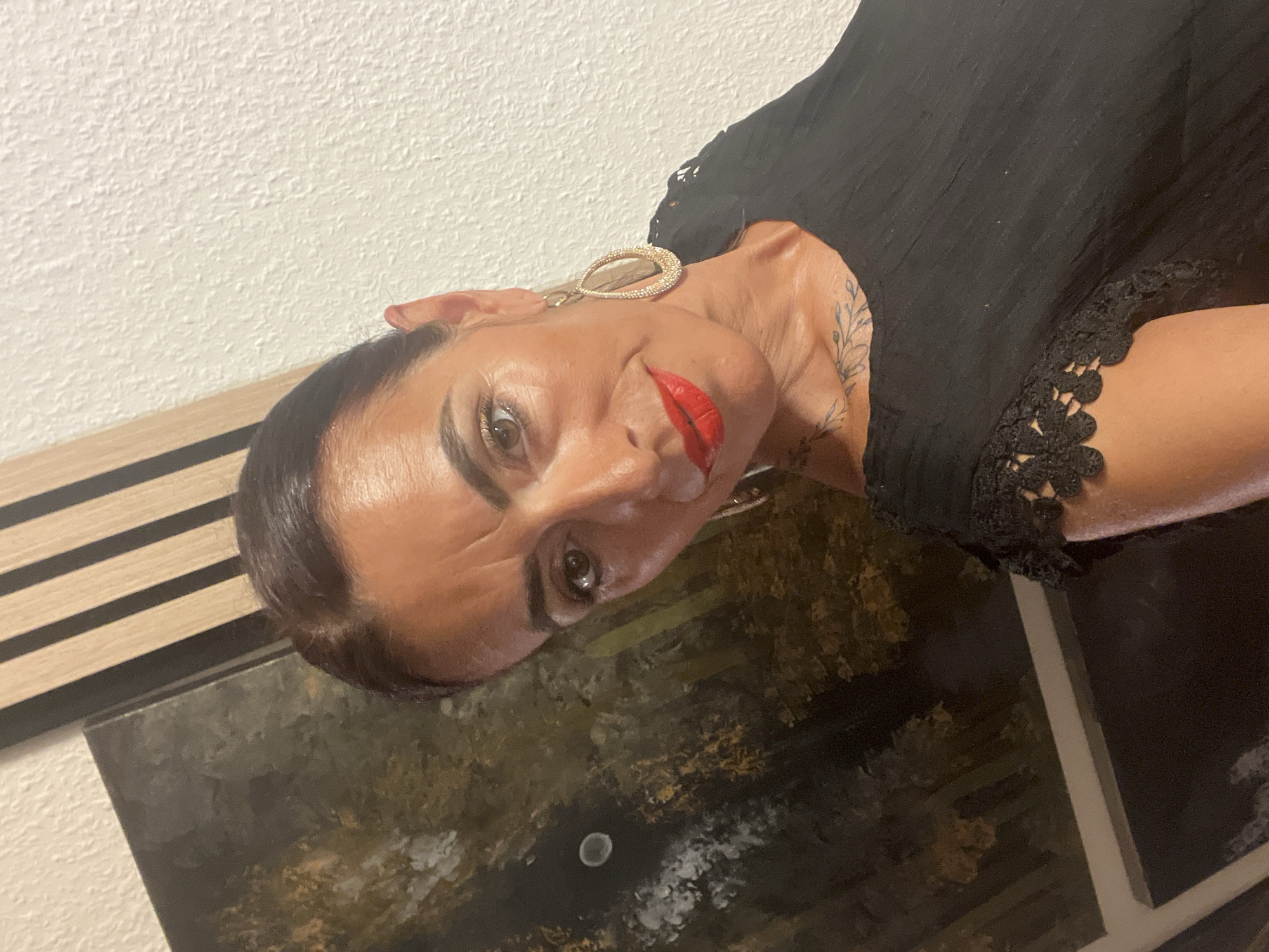 A woman with dark hair pulled back, brown eyes, and bright red lipstick poses for a selfie indoors. She is wearing a black top with lace detailing on the sleeves and large, gold, teardrop-shaped earrings. A tattoo of a floral design is visible on her neck and upper chest. Behind her, to the left, is a dark abstract painting with hints of gold and white. To the right of the painting is a section of wall with vertical wooden slats. The wall behind her to the right is textured and white. This image was taken in Konz, Germany.