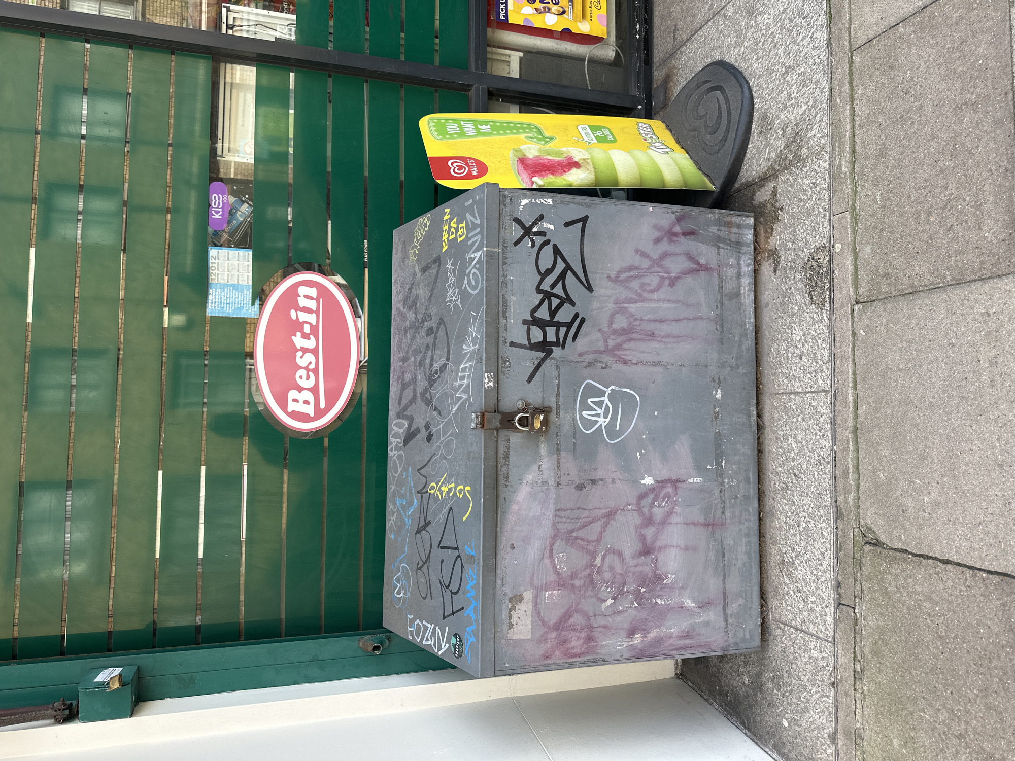 The scene depicts a street-level view in the City of London, United Kingdom, featuring a heavily graffitied metal utility box positioned against the closed security shutter of a commercial establishment. The primary subject is a dark grey, rectangular metal storage or utility box, approximately waist-high, resting on light grey pavement tiles. Its lid is secured by a small brass-colored padlock. The box is extensively marked with various graffiti tags in black, blue, yellow, and white, alongside faded maroon spray paint stains on its front and top surfaces.
Behind the box, a dark green corrugated metal roll-down security shutter covers a shop entrance. Through the horizontal slats of the shutter, elements of the shop interior and a brick building opposite are partially visible. A prominent red and white oval sign reading "Best-in" is affixed to the shutter, positioned above and to the right of the box. Nearby, a "KISS 100" sticker and other faded promotional posters are visible through the shutter's gaps.
To the immediate right of the metal box, a bright yellow and green "Wall's" ice cream standee, shaped like a "Twister" ice cream, advertises "Gluten Free" and "76 Calories." The standee features the Wall's heart logo and text reading "YOU WANT ME."
The ground consists of varied grey paving. The box sits on square, textured light grey tiles, which transition to larger, smoother concrete slabs in the foreground. A dark linear drain cover runs across the width of the pavement. No individuals are present in the frame.
