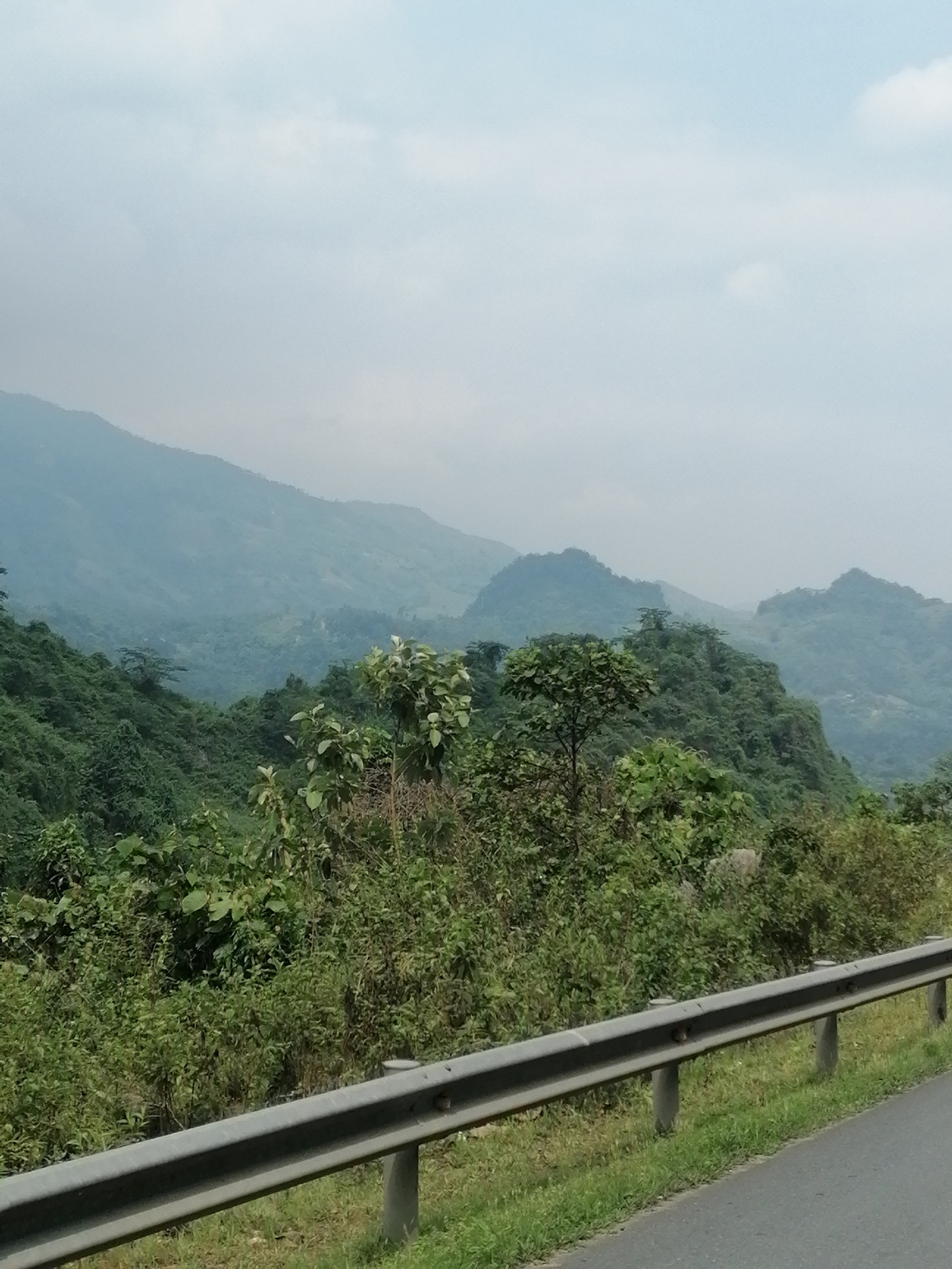 The image captures a mountainous landscape under an overcast sky. Lush green vegetation covers the hills and slopes. A gray guardrail lines the edge of a road in the foreground. The location is in the city of Unknown, Unknown.