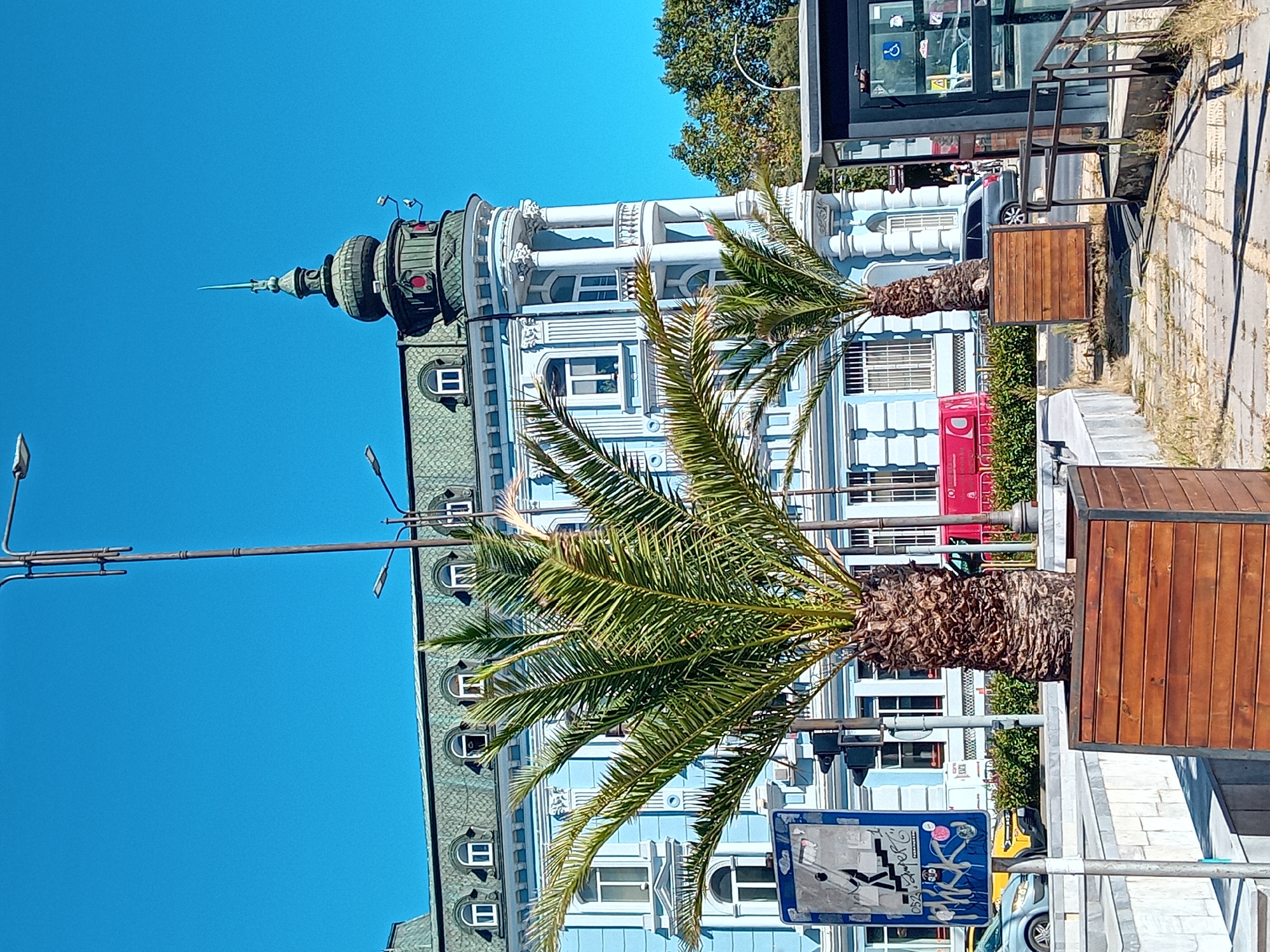 The image depicts the iconic "Synodal Palace" in Varna, Bulgaria, a striking example of Neo-Baroque architecture. The pale blue facade of the building, adorned with white columns and ornate windows, dominates the background. A distinctive green domed tower with a spire crowns the corner of the building, a prominent landmark of the city. In the foreground, two potted palm trees add a tropical touch to the urban setting, their fronds reaching towards the clear blue sky. A street sign with a symbol of stairs and a person is visible on the left, indicating pedestrian accessibility. A bus stop shelter with a glass enclosure is situated on the right, with railings and paved pathways. The scene is set on a sunny day, suggesting pleasant weather conditions. The image captures a vibrant and picturesque corner of Varna, showcasing its architectural heritage and urban amenities.