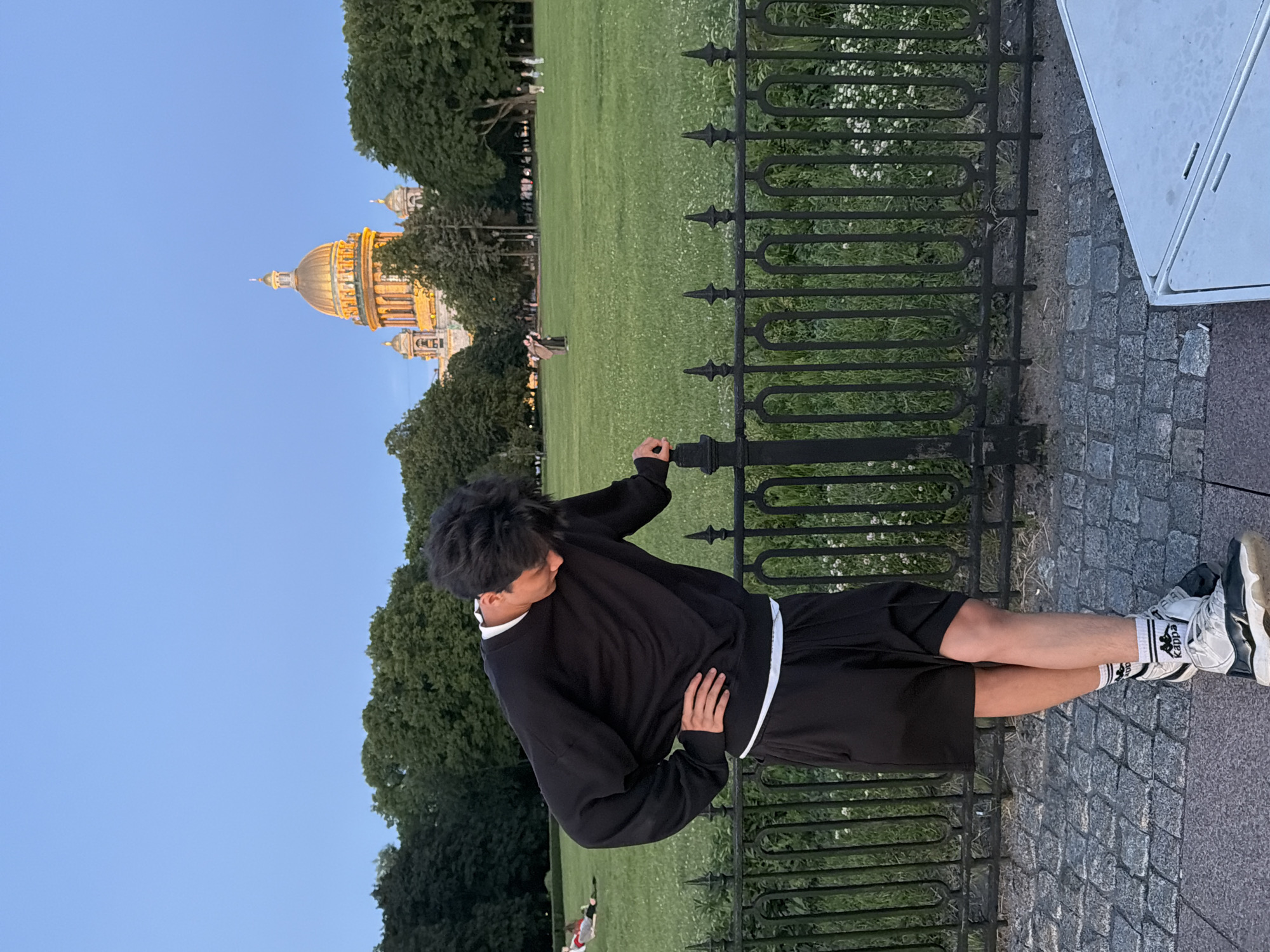 The media file captures a young person standing in an urban park with a large, domed building in the background, possibly St. Isaac's Cathedral in Saint Petersburg, Russia. The setting appears to be an outdoor public space with a well-maintained lawn, a decorative black metal fence, and cobblestone pavement. Trees line the perimeter of the park, and a bright blue sky suggests pleasant weather. The time of day is likely late afternoon or early evening, as indicated by the warm lighting on the building and the clear sky.
The primary subject is a young person dressed in black athletic wear, including a sweatshirt and shorts, with white and black socks and sneakers. They are leaning against the fence, with one hand on the fence and the other on their hip, looking away from the camera. Their posture suggests a relaxed or pensive mood. In the background, a few other people are scattered on the lawn, some appearing to be walking or sitting. The architectural style of the dome and building indicates a historical European city. The visible text on the person's socks reads "Kappa."