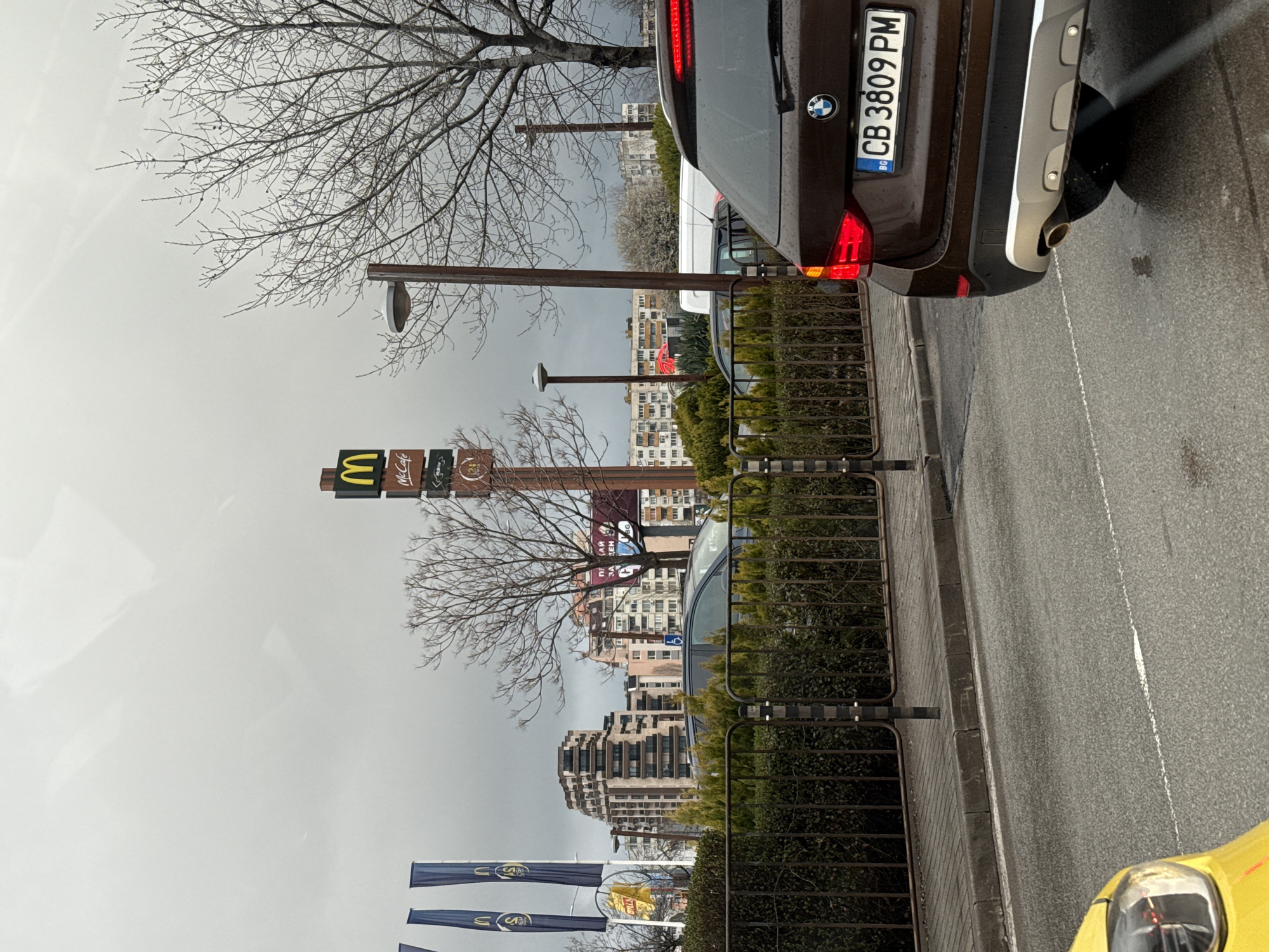 The image presents an urban scene in Sofia, Bulgaria, on an overcast day with a wet asphalt road, indicating recent precipitation. The perspective is from a vehicle, with the yellow headlight and hood of a car visible in the lower left foreground. A brown BMW SUV, bearing Bulgarian license plate "CB 3809 PM" with illuminated red taillights, is positioned in the mid-right foreground, facing away from the viewer. Its rear bumper features a silver-colored lower trim and a visible exhaust pipe.
A black metal fence, interspersed with black and white striped bollards, separates the roadway from a landscaped area containing trimmed green bushes and multiple bare deciduous trees, suggesting a cooler season. A tall brown pole sign for McDonald's stands prominently in the midground, displaying the yellow 'M' logo, "McCafé," and "24H" branding. To the left of the sign, three dark blue advertising flags with yellow McDonald's logos are visible. Behind the landscaping, other vehicles are partially visible, including a dark minivan with a blue disabled parking symbol displayed on its windshield and a white van. Tall brown streetlamp poles with gray, disk-shaped fixtures are also present.
The background features a dense urban environment beneath an entirely overcast, light gray sky. A distinctive multi-story modern building with a terraced, stacked facade and numerous balconies is visible in the left-center background. Various other multi-story residential or commercial buildings, exhibiting different architectural styles and colors, extend across the background. A large red and white billboard displaying "ПРАЙМ ЗАСТРАХОВАНЕ" (Prime Insurance) in Cyrillic is partially visible among the background buildings and trees. No individuals are visible within the frame. This scene is located in the vicinity of Bulgaria Boulevard and Todor Kableshkov Street in Sofia, specifically near the McDonald's at Bulgaria Blvd 118, 1404 kv. Krastova vada.